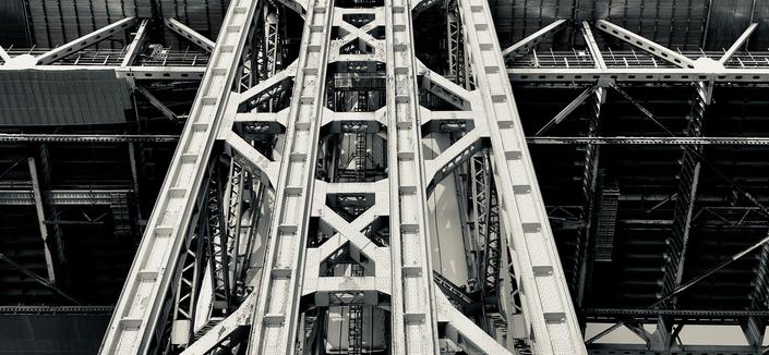 A noir photograph in letterbox format of the monumental structural framework of the George Washington Bridge on a sunny Autumn morning. The view is from directly underneath, looking straight up into the towering lattice of the massive steel column. Visible within the structure are maintenance ladders. The bridge fills the entire frame.
