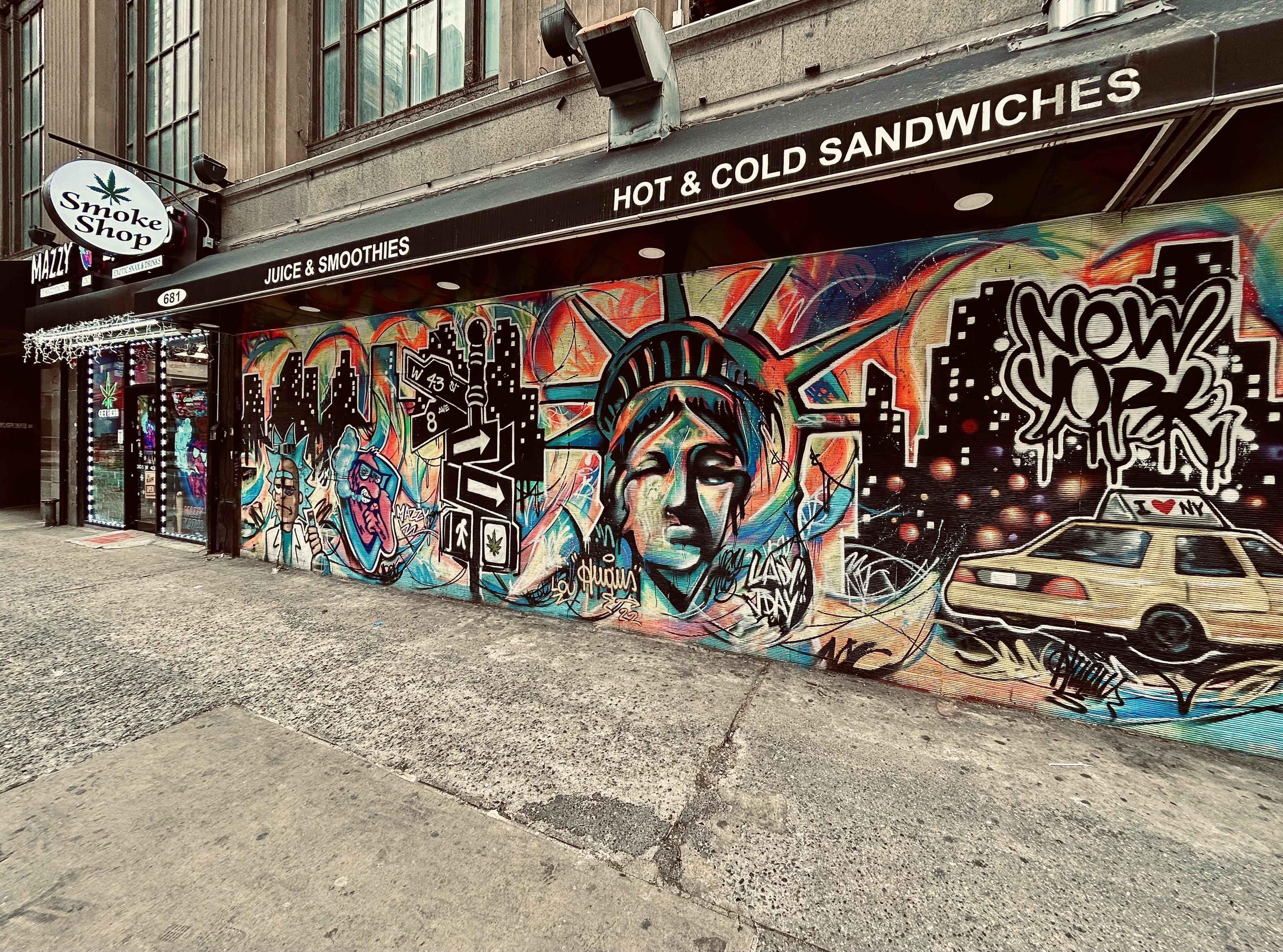 A color photograph of the street scene along 44th Street at the intersection with Eighth Avenue on an overcast winter morning. The focal point of the image is a street mural that dominates the facade of an exterior wall for a cannabis dispensary. The mural is a densely packed, colorful composition featuring iconic New York City imagery rendered in a dynamic mix of graffiti, pop art, and illustration. The central focus is a large, stylized, and slightly distressed face of the Statue of Liberty, rendered in blue, green, and flesh tones, wearing her crown. To her right, a speeding yellow New York City taxi cab is depicted driving across a gritty, black cityscape silhouette with twinkling lights. Bold, black-outlined graffiti tags and text overlays, including one reading "NEW YORK," are scattered across the composition. The mural covers the lower portion of the building's exterior, running beneath a dark awning that prominently features white text reading "HOT & COLD SANDWICHES" and "JUICE & SMOOTHIES." To the far left, the mural stops at the entrance of a business with a small white circular, back-lit sign featuring a cannabis leaf icon and the words “Smoke Shop.” The ground in the foreground is a rough, concrete sidewalk.