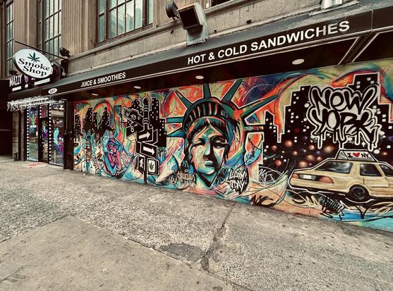 A color photograph of the street scene along 44th Street at the intersection with Eighth Avenue on an overcast winter morning. The focal point of the image is a street mural that dominates the facade of an exterior wall for a cannabis dispensary. The mural is a densely packed, colorful composition featuring iconic New York City imagery rendered in a dynamic mix of graffiti, pop art, and illustration. The central focus is a large, stylized, and slightly distressed face of the Statue of Liberty, rendered in blue, green, and flesh tones, wearing her crown. To her right, a speeding yellow New York City taxi cab is depicted driving across a gritty, black cityscape silhouette with twinkling lights. Bold, black-outlined graffiti tags and text overlays, including one reading "NEW YORK," are scattered across the composition. The mural covers the lower portion of the building's exterior, running beneath a dark awning that prominently features white text reading "HOT & COLD SANDWICHES" and "JUICE & SMOOTHIES." To the far left, the mural stops at the entrance of a business with a small white circular, back-lit sign featuring a cannabis leaf icon and the words “Smoke Shop.” The ground in the foreground is a rough, concrete sidewalk.