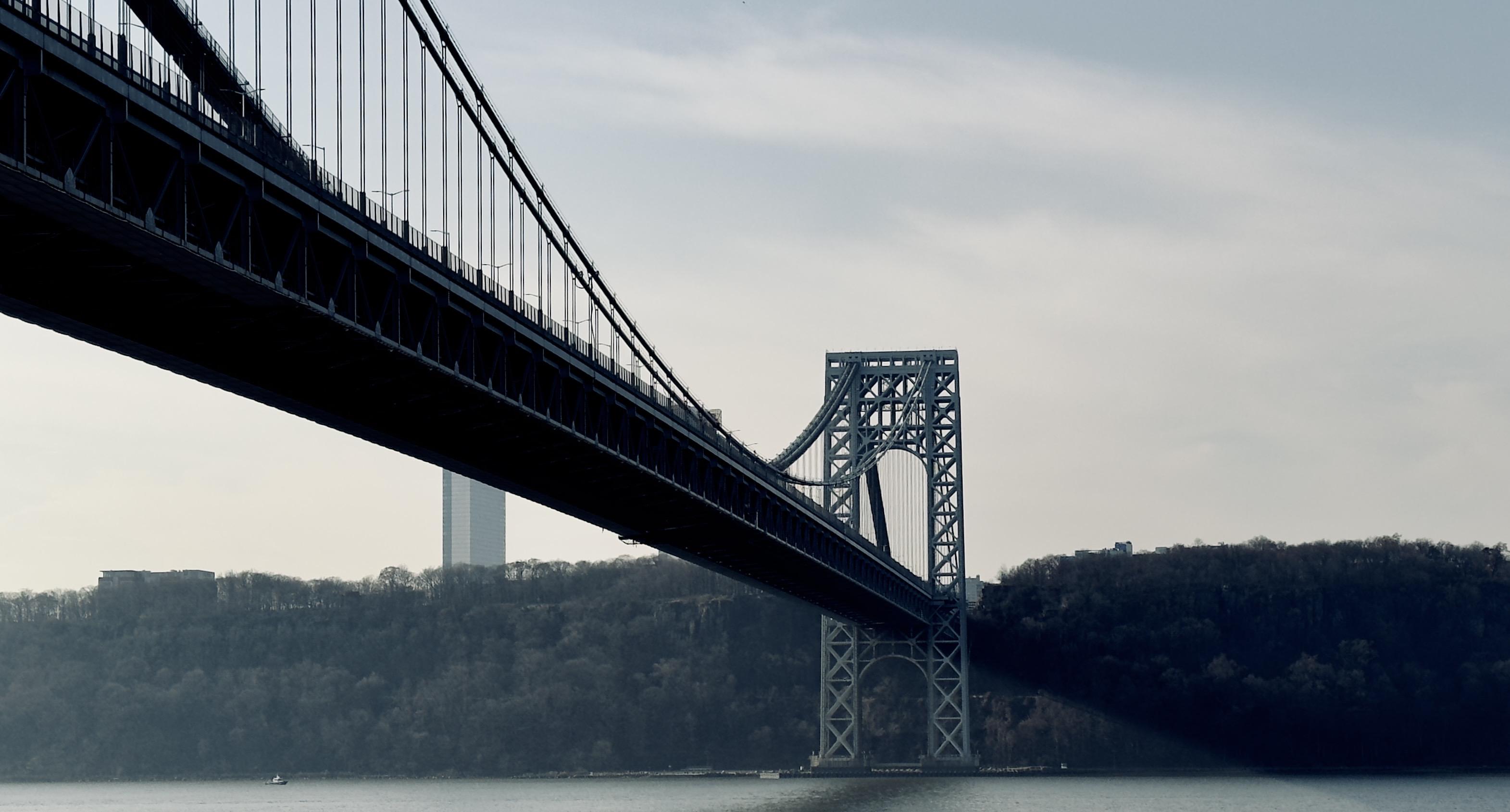 A color photograph shot from a low angle of the George Washington Bridge, spanning the Hudson River, captured on an overcast afternoon. The massive, dark steel structure of the main suspension span dominates the upper left and center of the frame, angled diagonally from the top-left corner down toward the center-right. The suspension cables are clearly visible, rising toward the unseen roadway above. In the distance behind the bridge tower, a single, tall, slender skyscraper is faintly visible against the hazy sky. The foreground is the calm, dark water of the river, with a small, white vessel barely visible near the bottom left edge of the frame. The lighting is diffused and cool, giving the overall image a muted tone.