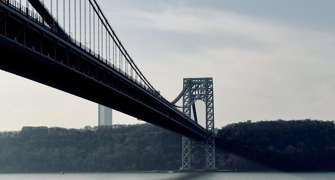 A color photograph shot from a low angle of the George Washington Bridge, spanning the Hudson River, captured on an overcast afternoon. The massive, dark steel structure of the main suspension span dominates the upper left and center of the frame, angled diagonally from the top-left corner down toward the center-right. The suspension cables are clearly visible, rising toward the unseen roadway above. In the distance behind the bridge tower, a single, tall, slender skyscraper is faintly visible against the hazy sky. The foreground is the calm, dark water of the river, with a small, white vessel barely visible near the bottom left edge of the frame. The lighting is diffused and cool, giving the overall image a muted tone.