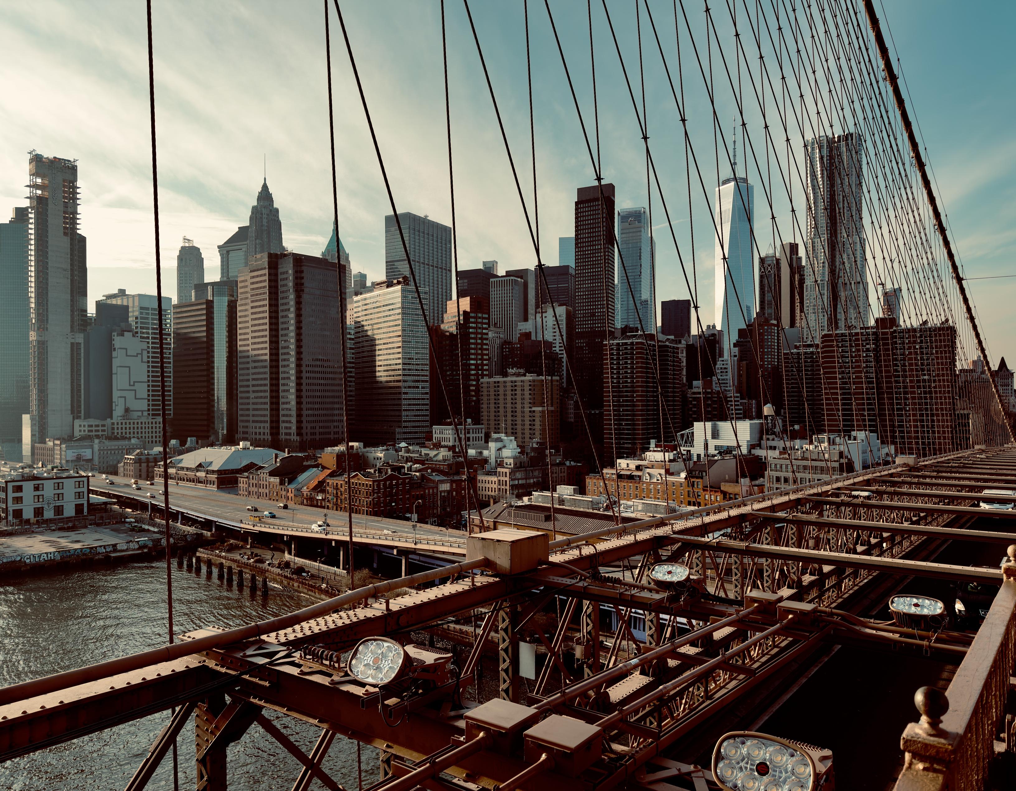 A color, low-angle photograph taken from the Brooklyn Bridge walkway, looking toward the Lower Manhattan skyline late on a mostly clear, late winter afternoon. The foreground is dominated by the bridge's intricate, dark brown metal truss structure and several of the thick vertical suspension cables that stretch diagonally across the top right of the frame. The metallic structure has several circular lighting fixtures attached to the beams. To the left, the East River's dark water meets the land, where an elevated highway with moderate traffic passes below the skyline. The warm afternoon sun casts a deep, saturated tone over the scene. The prominent Lower Manhattan skyline fills the middle and background with a dense collection of modernist and historic skyscrapers. The buildings are primarily composed of dark glass and brick. Key landmarks identifiable include One World Trade Center, which rises noticeably above the rest of the buildings. The ambient lighting suggests the sun is low on the horizon, creating long, subtle shadows and giving the entire image a rich, slightly moody, golden-brown hue that emphasizes the cold weather and the scale of the urban environment.