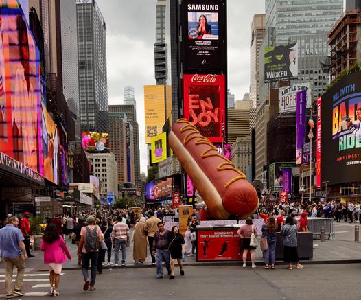 A color photograph of the street scene in a crowded section of Times Square in New York City on a cloudy summer morning day. The focal point of the image is a massive art installation designed to look like a hot dog. The colossal hot dog, complete with a wavy line of yellow mustard, rests on the ground at the center-right of the frame. It occupies a significant portion of the lower foreground and mid-ground, making the people nearby appear small in comparison. Behind the installation, the street is densely populated with pedestrians, and the backdrop is defined by the many towering buildings covered in bright, illuminated digital billboards displaying advertisements for various brands, shows like "Wicked," and a large Coca-Cola and Samsung screen at the top center. People are walking in all directions, some looking toward the hot dog installation, and others moving past it. The overall color palette is dominated by the bright, saturated colors of the digital screens and contrasting with the neutral tones of the concrete streets, cloudy sky, and the glass and steel of the skyscrapers.