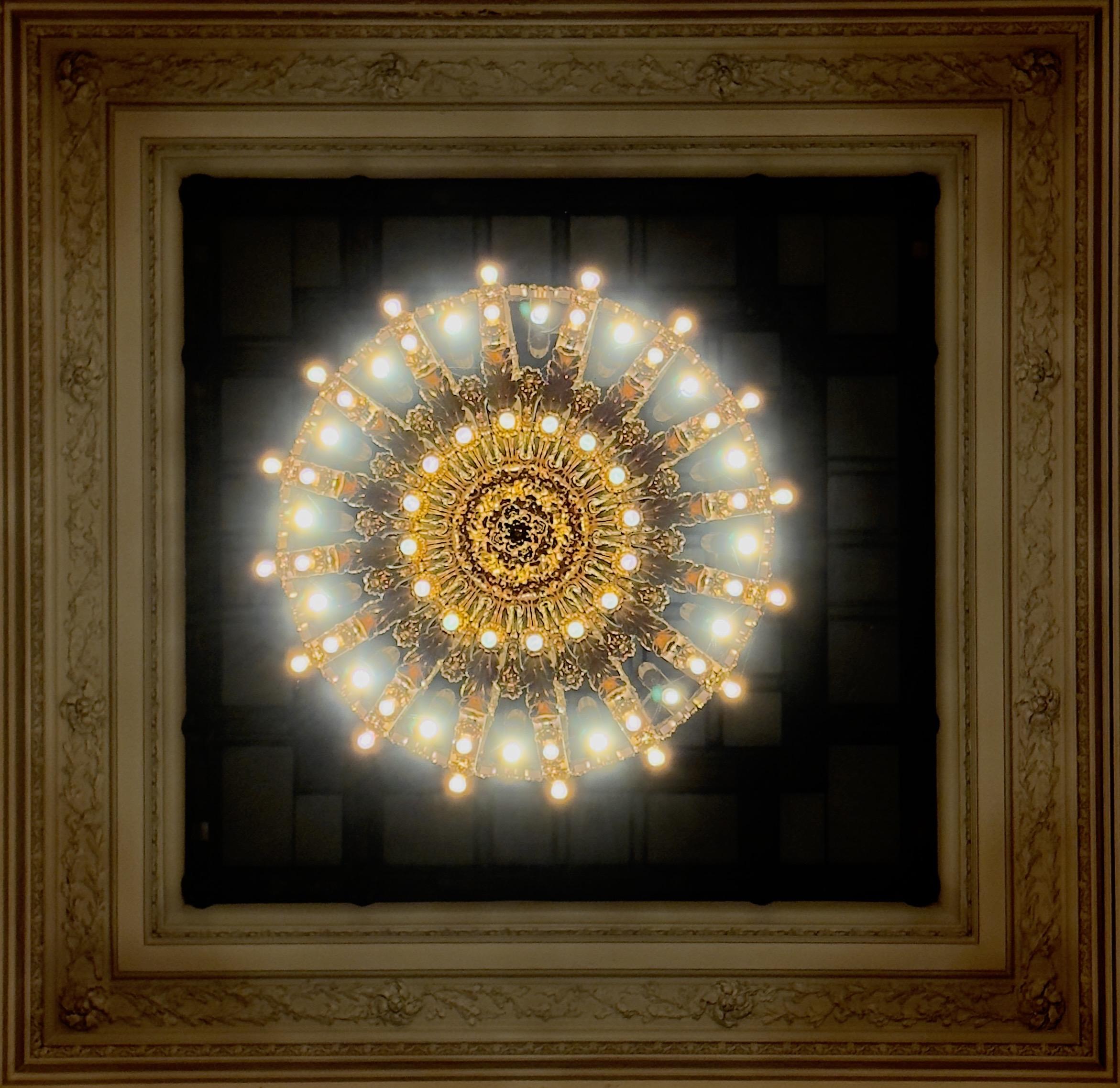 A color photograph taken looking up at a large, ornate, circular chandelier centered in the middle of a coffered ceiling panel located in Grand Central Terminal, New York City. The chandelier has a complex, multi-tiered brass and gold-toned frame and is encircled by numerous exposed, glowing incandescent bulbs, creating a strong, bright, star-like effect with light rays shining outwards. The circular light fixture is surrounded by a square, decorative stone ceiling border, which is framed by a wider, lighter-toned square molding featuring intricate relief details of floral and acorn motifs, all against a dark, recessed background.