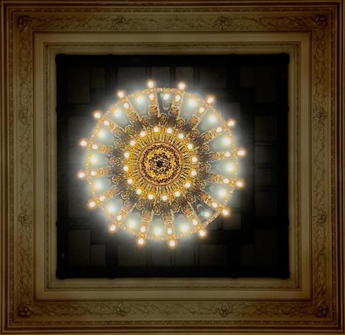 A color photograph taken looking up at a large, ornate, circular chandelier centered in the middle of a coffered ceiling panel located in Grand Central Terminal, New York City. The chandelier has a complex, multi-tiered brass and gold-toned frame and is encircled by numerous exposed, glowing incandescent bulbs, creating a strong, bright, star-like effect with light rays shining outwards. The circular light fixture is surrounded by a square, decorative stone ceiling border, which is framed by a wider, lighter-toned square molding featuring intricate relief details of floral and acorn motifs, all against a dark, recessed background.