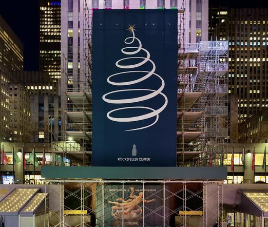 A color photograph of the Rockefeller Center Christmas tree setup this year, prior to being lit on December 3rd. It is nighttime. The focal point of the image is a massive vertical dark blue banner hanging centrally over a complex metal scaffolding structure that has been built around the tree, and obscuring the usual view of the Rockefeller Center plaza backdrop. The banner features a large, stylized white line drawing of a Christmas tree formed by a continuous, tapering spiral, topped with a gold starburst design. At the bottom of the banner, the Art Deco "Rockefeller Center" logo is printed in white. The scaffolding extends high into the air, flanked by the illuminated cool-blue and yellow windows of towering city skyscrapers at night. Below the main blue banner and a green construction ledge, a lower barrier displays a graphic of the famous gold Prometheus statue with text reading "Restoring a Masterpiece" and "The Rink." The plaza level is lined with flags on poles, and small tent-like structures with peaked roofs and warm string lights frame the bottom corners of the scene.