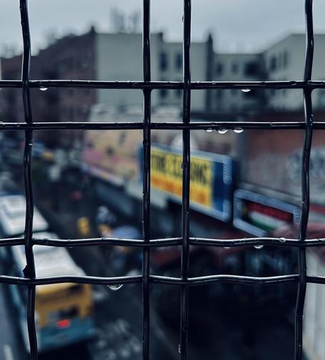 A color photograph, taken up-close and looking through a dark, grid-patterned metal fence that encloses a windowed view from the elevated train platform at Fordham Road Station in the Bronx. Tar scene is captured on a rainy day. The fence, which fills the foreground, is the sharpest element in the image, with several glistening water droplets clinging to the thick vertical and horizontal wires. This sharp foreground contrasts heavily with the soft, shallow depth of field in the background, which is blurred by the focus. The blurred background shows a street scene below and beyond. The left side features the top of a yellow and blue city bus a red tail light visible. On the right, a row of brick apartment buildings is visible behind a collection of large, brightly colored commercial signs atop stores along the street. The most prominent sign is a long, horizontal one with red, yellow, and blue text. This adds a pop of vibrant, blurry strokes of color to the otherwise muted, overcast palette of greys and browns. The overall mood is cool, damp, and urban.