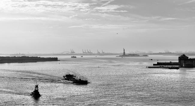 A noir, wide-angle photograph of the Upper New York Bay on a slightly hazy day. In the immediate foreground, the textured water is disrupted by two vessels. A large tanker ship is the most prominent element, moving across the frame toward the center-right, leaving a clear wake. A smaller vessel is positioned closer to the bottom-left edge. The Statue of Liberty is clearly visible in the midground, silhouetted against the bright, hazy sky on her island base. To the right, a large, dark angular docking facility juts into the water. The distant horizon is dominated by an industrial landscape where dozens of tall, distinct gantry cranes line the far shore. These structures are partially obscured by the atmospheric haze, contrasting sharply with the bright sky that contains a few scattered clouds and subtle contrails overhead.