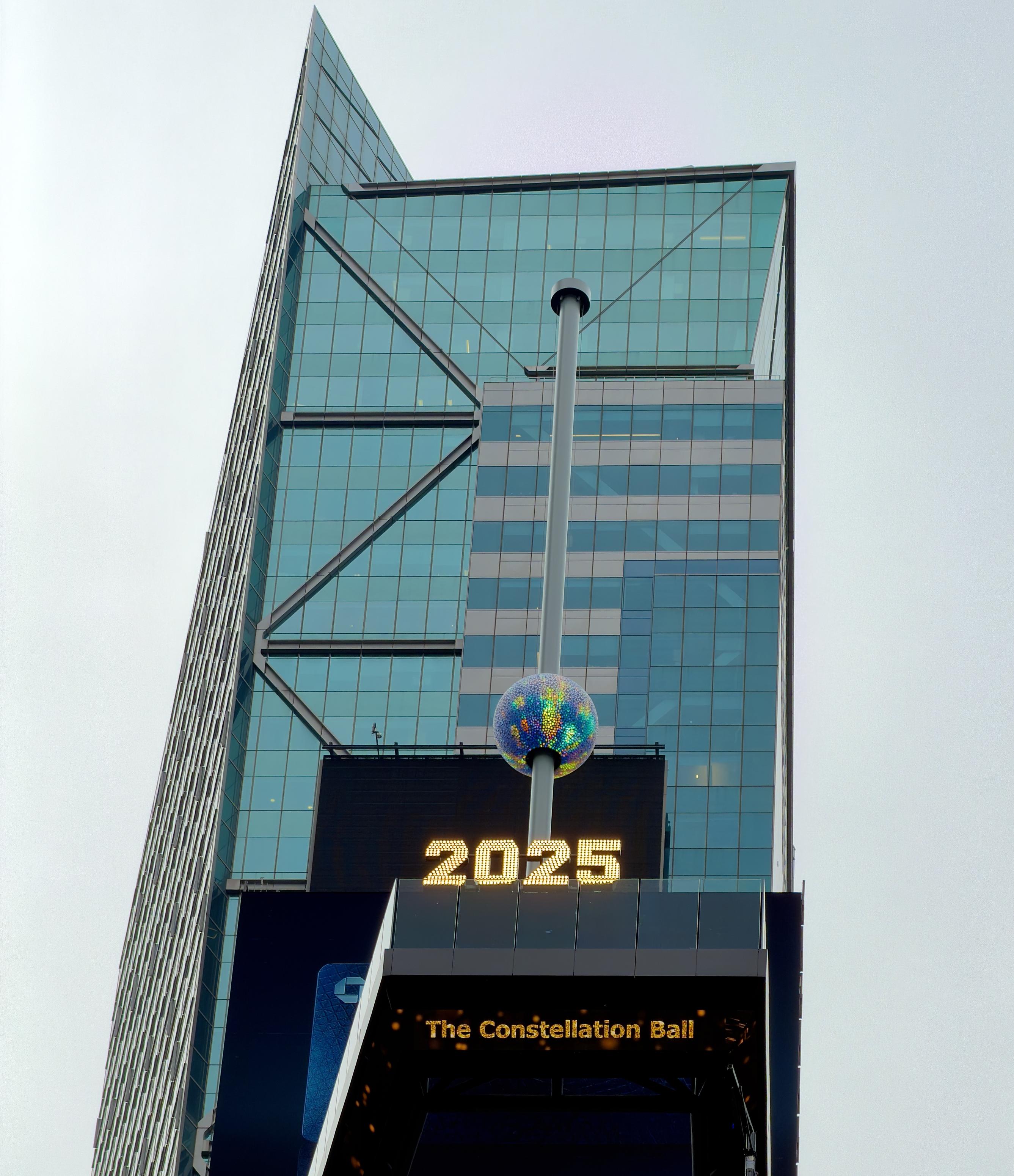 A color, low-angle, portrait orientation photograph of the new Times Square New Year’s Eve ball installation against the backdrop of a modern skyscraper and an overcast sky. The ball itself is the centerpiece of the image. It is made up of a glittering geodesic sphere covered in numerous circular, light-refracting crystals that give it a multicolored, galaxy-like appearance. The sphere is positioned near the bottom of a tall, silver pole that extends straight up. Directly beneath the ball and pole is a dark electronic sign displaying the bright, amber-yellow numerals "2025," an older sign awaiting replacement for the upcoming year, and a smaller, yellow text sign below it that reads, "The Constellation Ball." The surrounding architecture is dominated by a sharply angled glass facade on the left, featuring visible diagonal structural cross-beams, and a less reflective, multi-story structure on the right.