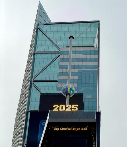 A color, low-angle, portrait orientation photograph of the new Times Square New Year’s Eve ball installation against the backdrop of a modern skyscraper and an overcast sky. The ball itself is the centerpiece of the image. It is made up of a glittering geodesic sphere covered in numerous circular, light-refracting crystals that give it a multicolored, galaxy-like appearance. The sphere is positioned near the bottom of a tall, silver pole that extends straight up. Directly beneath the ball and pole is a dark electronic sign displaying the bright, amber-yellow numerals "2025," an older sign awaiting replacement for the upcoming year, and a smaller, yellow text sign below it that reads, "The Constellation Ball." The surrounding architecture is dominated by a sharply angled glass facade on the left, featuring visible diagonal structural cross-beams, and a less reflective, multi-story structure on the right.