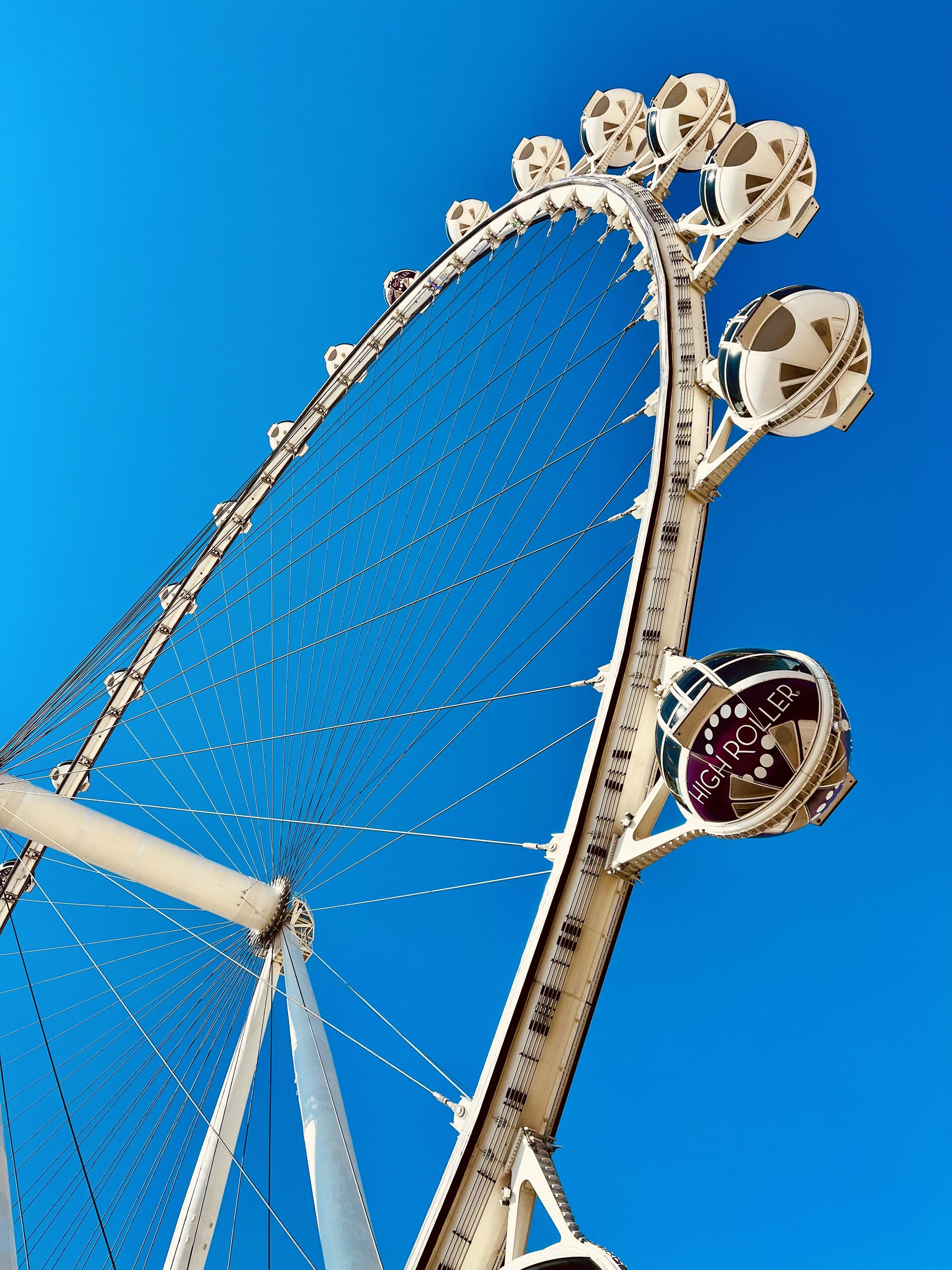 A color, low-angle photograph that captures the massive, white structural spine and cables of the High Roller Wheel 🎡 along The Strip in Las Vegas. It is silhouetted against a brilliant and cloudless deep blue sky. The wheel dominates the frame, curving sharply upward from the bottom left toward the upper right from a perspective that emphasizes its immense scale. A prominent, dark-tinted passenger gondola, clearly marked with the purple and white "HIGH ROLLER" logo, is visible in the foreground attached to the main support structure on the right. The scene is highly saturated and warm.