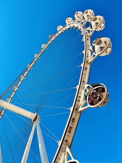 A color, low-angle photograph that captures the massive, white structural spine and cables of the High Roller Wheel 🎡 along The Strip in Las Vegas. It is silhouetted against a brilliant and cloudless deep blue sky. The wheel dominates the frame, curving sharply upward from the bottom left toward the upper right from a perspective that emphasizes its immense scale. A prominent, dark-tinted passenger gondola, clearly marked with the purple and white "HIGH ROLLER" logo, is visible in the foreground attached to the main support structure on the right. The scene is highly saturated and warm.