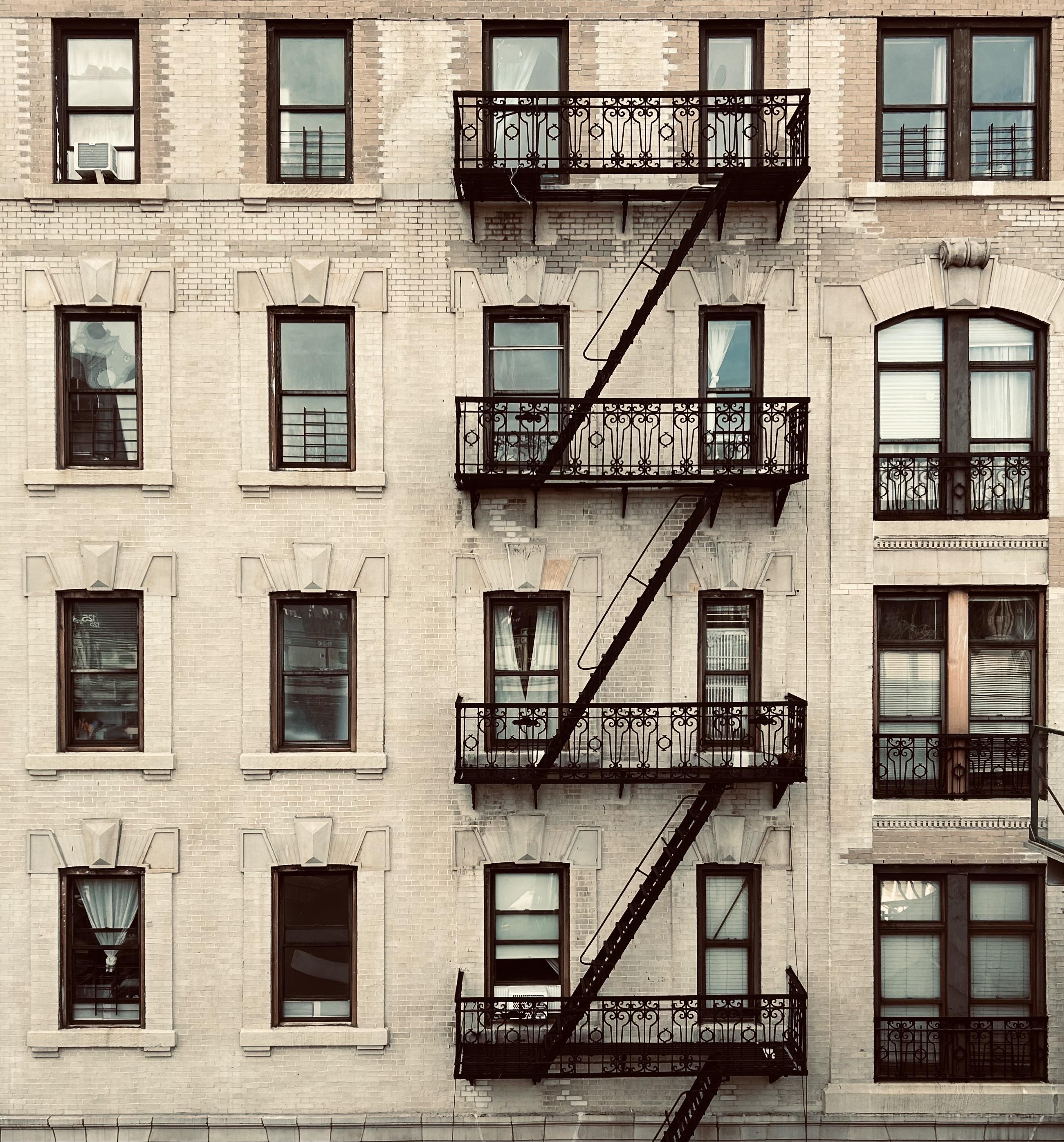 A color, desaturated photograph of the exterior of an apartment building along Broadway in the Harlem neighborhood of Manhattan. The facade is constructed of light, cream colored brick with darker mortar, and the image is dominated by a grid of vertically stacked windows and a prominent black iron fire escape running diagonally across the right side. The windows have various treatments: some are simple sash windows with black frames, others have black metal security bars, and some are adorned with small, ornate iron window guards that match the style of the fire escape balconies. On the far right, the windows are larger and more varied in shape, including a prominent arched window on the third floor. The centerpiece of the composition is the rusted, wrought iron fire escape, featuring highly decorative, detailed balustrades on each of the four balconies, contrasting strongly with the pale brickwork. The ladders connecting the balconies run steeply down and to the left, which creates a zigzag pattern. The overall tone is gritty yet elegant, capturing the classic pre-war architectural style and urban characteristics. The lighting is relatively flat on a partly overcast morning, enhancing the textural details of the brick and the decorative ironwork.