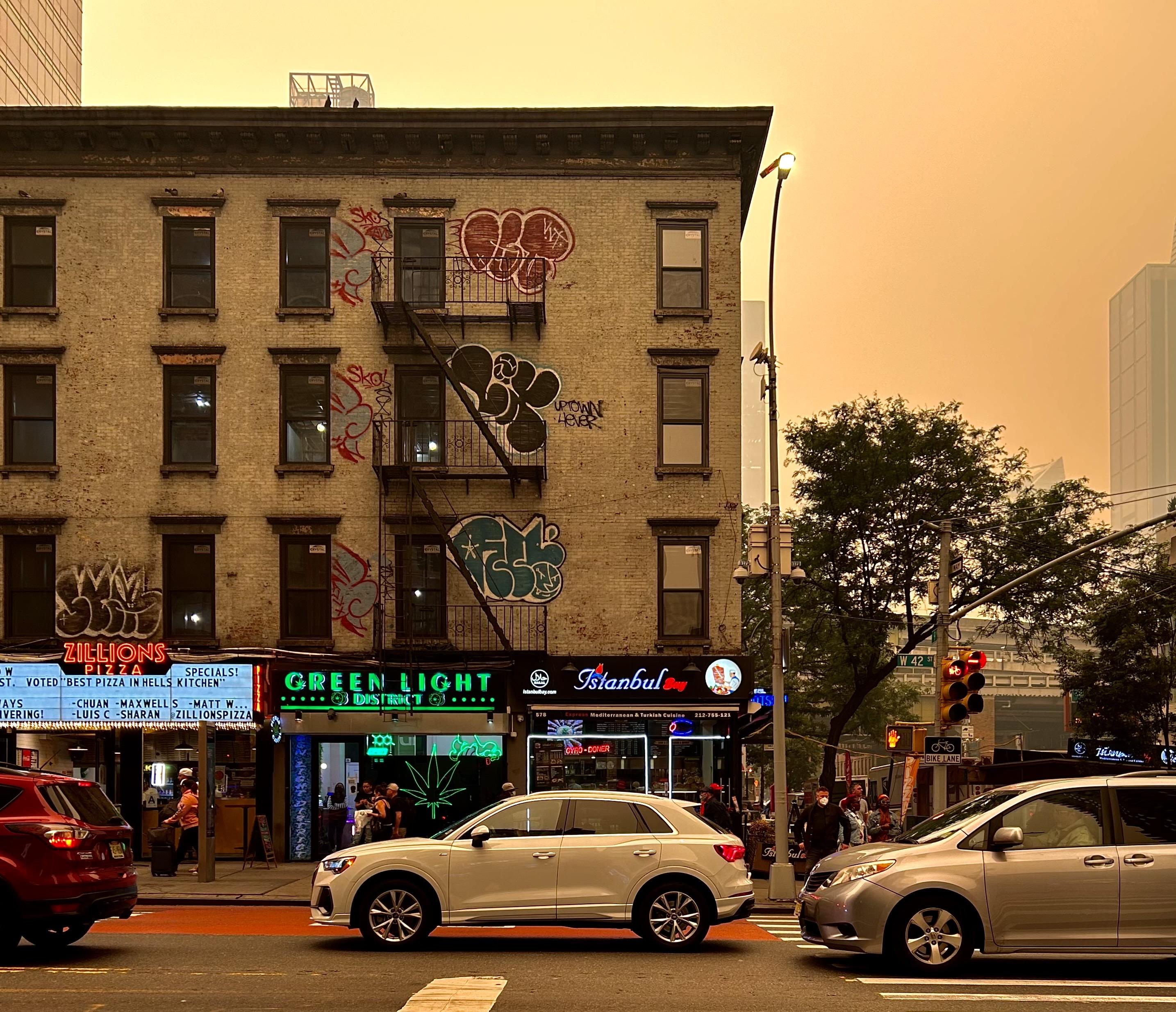 A color photograph in square aspect ratio that captures the street scene in Manhattan on the early afternoon June 7, 2023, during the severe air quality crisis caused by smoke from Canadian wildfires. The image is dominated by an apocalyptic, heavy sepia toned smog that saturates the sky and the overall lighting. In the center, a four-story brick apartment building with several fire escapes, heavily adorned with large, black and blue graffiti tags. At street level, the building is lined with neon-lit storefronts, including "Zillions Pizza," "Green Light dispensary," and "Istanbul Turkish cuisine," with a small crowd of pedestrians visible near the entrances. The dispensary has a large neon cannabis leaf inside a window. The street is busy with vehicular traffic, featuring a silver minivan, a white SUV, and a red SUV. The pervasive orange light from the thick smoke transforms the typically vibrant cityscape into a muted, almost monochromatic scene. The bright neon signs contrast sharply with the dark, gritty texture of the old brick building and the uniform haze overhead.