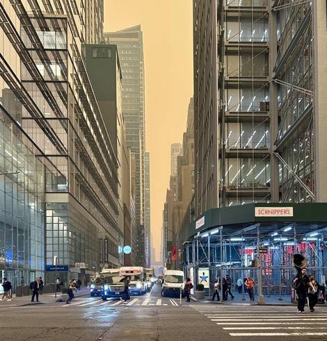 A color photograph taken on 41st Street at the intersection with Eighth Avenue in Manhattan, during the Canadian wildfire event on June 7th, 2023. The sky above the towering glass and steel skyscrapers is completely obscured by an exceptionally thick, smoky haze, which casts an ominous, deep sepia-orange glow over the entire scene. The orange light diffuses the urban setting, slightly muting the colors of the modern buildings that flank the street on both sides, and significantly reducing visibility down the avenue's canyon toward the background. Pedestrians are visible crossing the wide intersection, and several vehicles, including a van and a transit-style bus, are positioned near the crosswalk. Most notably, on the far right side of the crosswalk, a man carrying a large, shiny brass sousaphone is crossing the street.