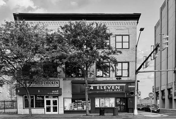 A noir, wide photograph of the street scene along Atlantic Avenue in Atlantic City, New Jersey. The focal point of the scene is two commercial storefronts on the ground floor of a three-story brick building. On the left is a restaurant named Crown Fried Chicken, with a sign that advertises halal chicken. The entrance is a glass door underneath a dark awning. To the right of the restaurant is a convenience store named 4 Eleven Food Mart. Both storefronts have large windows displaying promotional materials, and a trash receptacle, and recycling receptacle sit between the two entrances on the sidewalk. Two mature deciduous trees with full foliage stand on the narrow sidewalk in front of the building, partially obscuring the upper floors of the building. The upper two stories of the building are constructed with light-colored brick, featuring a row of five square multi-pane windows on the second and third floors. The building has a flat, dark roofline. To the right of the building, a street intersection is visible, marked by a traffic light and a tall street lamp post. Further right and slightly behind the storefront building, the side of a modern, multi-story building with a plain vertical facade dominates the background. A dark sedan is parked on the street near a 'DO NOT ENTER' sign in the distance on the far right. The sky is brought with scattered clouds defining the horizon.
