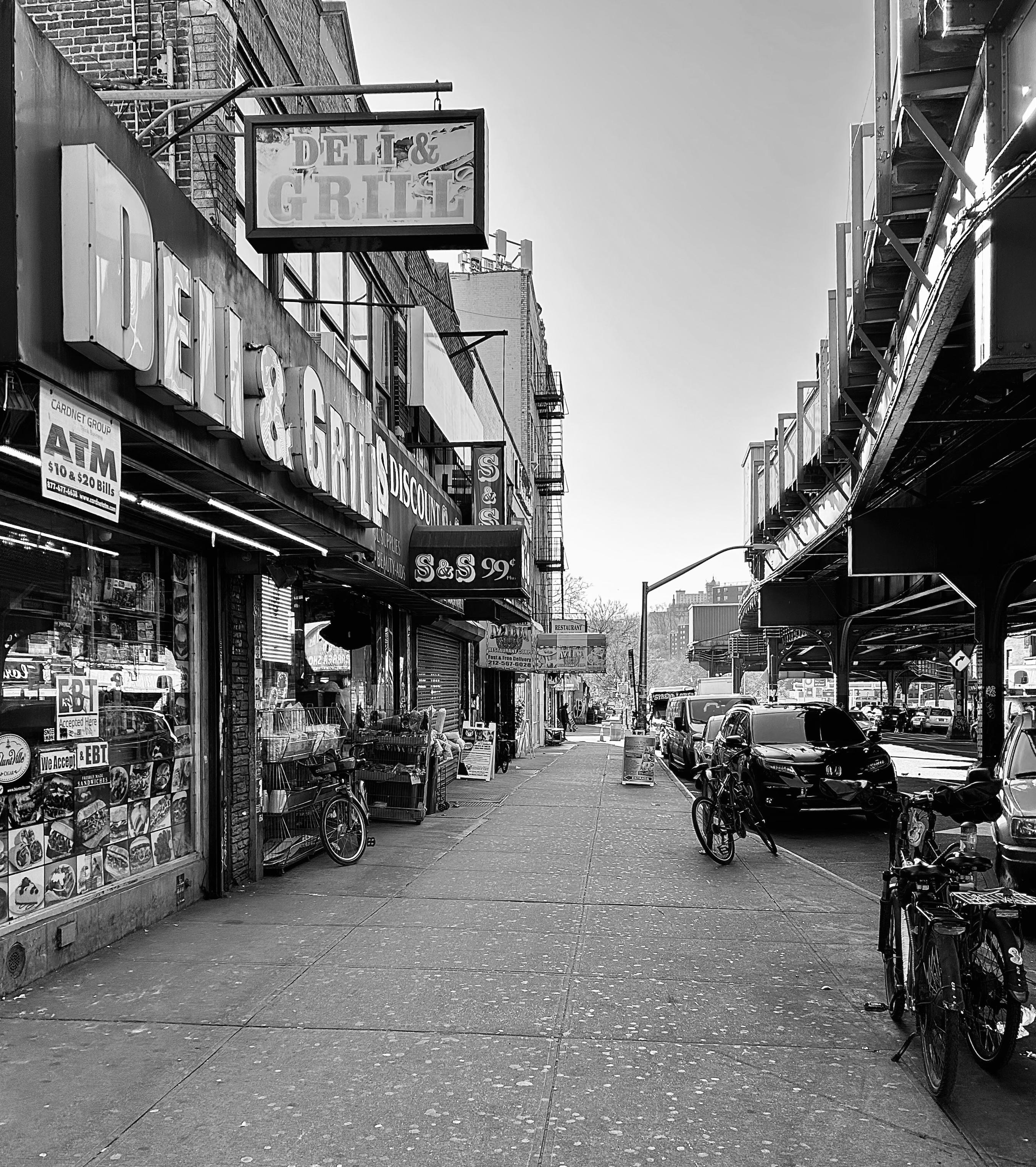 A noir, high-contrast photograph captures the street scene along Tenth Avenue in the Inwood neighborhood of Manhattan. On the left, a building facade prominently features large, bold signs for a "DELI & GRILL". The storefront windows are cluttered with various signs, product advertisements, and stickers, including one for an ATM and another indicating acceptance of EBT. Merchandise is visible inside the store and partially displayed to the sidewalk outside. On the right side of the sidewalk, several bicycles are parked near the storefront, and the paved sidewalk extends into the distance, narrowing the perspective. The right side of the image is dominated by the elevated subway line at 207th Street Station, which creates a deep shadow over the street and parked cars beneath it. The elevated track structure adds a strong linear element and industrial feel to the composition. Beyond the parked vehicles and the immediate urban architecture, the background shows a hint of distant buildings and a clear, bright sky.