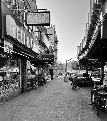 A noir, high-contrast photograph captures the street scene along Tenth Avenue in the Inwood neighborhood of Manhattan. On the left, a building facade prominently features large, bold signs for a "DELI & GRILL". The storefront windows are cluttered with various signs, product advertisements, and stickers, including one for an ATM and another indicating acceptance of EBT. Merchandise is visible inside the store and partially displayed to the sidewalk outside. On the right side of the sidewalk, several bicycles are parked near the storefront, and the paved sidewalk extends into the distance, narrowing the perspective. The right side of the image is dominated by the elevated subway line at 207th Street Station, which creates a deep shadow over the street and parked cars beneath it. The elevated track structure adds a strong linear element and industrial feel to the composition. Beyond the parked vehicles and the immediate urban architecture, the background shows a hint of distant buildings and a clear, bright sky.