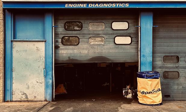 A color photograph in landscape orientation captures the partially opened entrance to an auto repair garage in Hempstead on Long Island, New York near sunset. The dominant colors are muted blues, grays, and browns for the slightly weathered industrial scene. The top of the image features a bright blue header spanning the width of the entrance with the text "ENGINE DIAGNOSTICS" clearly visible in yellow lettering. The entrance itself is composed of three main vertical sections: a metal side door on the left, a central, partially rolled-up metal garage door, and a closed section of a metal garage door on the right. The side door on the far left is an off-white metal, and notably, the word "LOVE" and a small heart shape are scratched onto its surface. The central garage door is a corrugated gray metal with a pattern of six horizontal oval windows across its upper half. The bottom half of this door is rolled up, revealing the darkened entrance where a few tools and objects are dimly visible on the concrete floor. To the right, against the closed section of the garage door, is a large, crinkled yellow industrial bag with the word "GOODYEAR" partially legible on it, contrasting with the surrounding blues and grays.