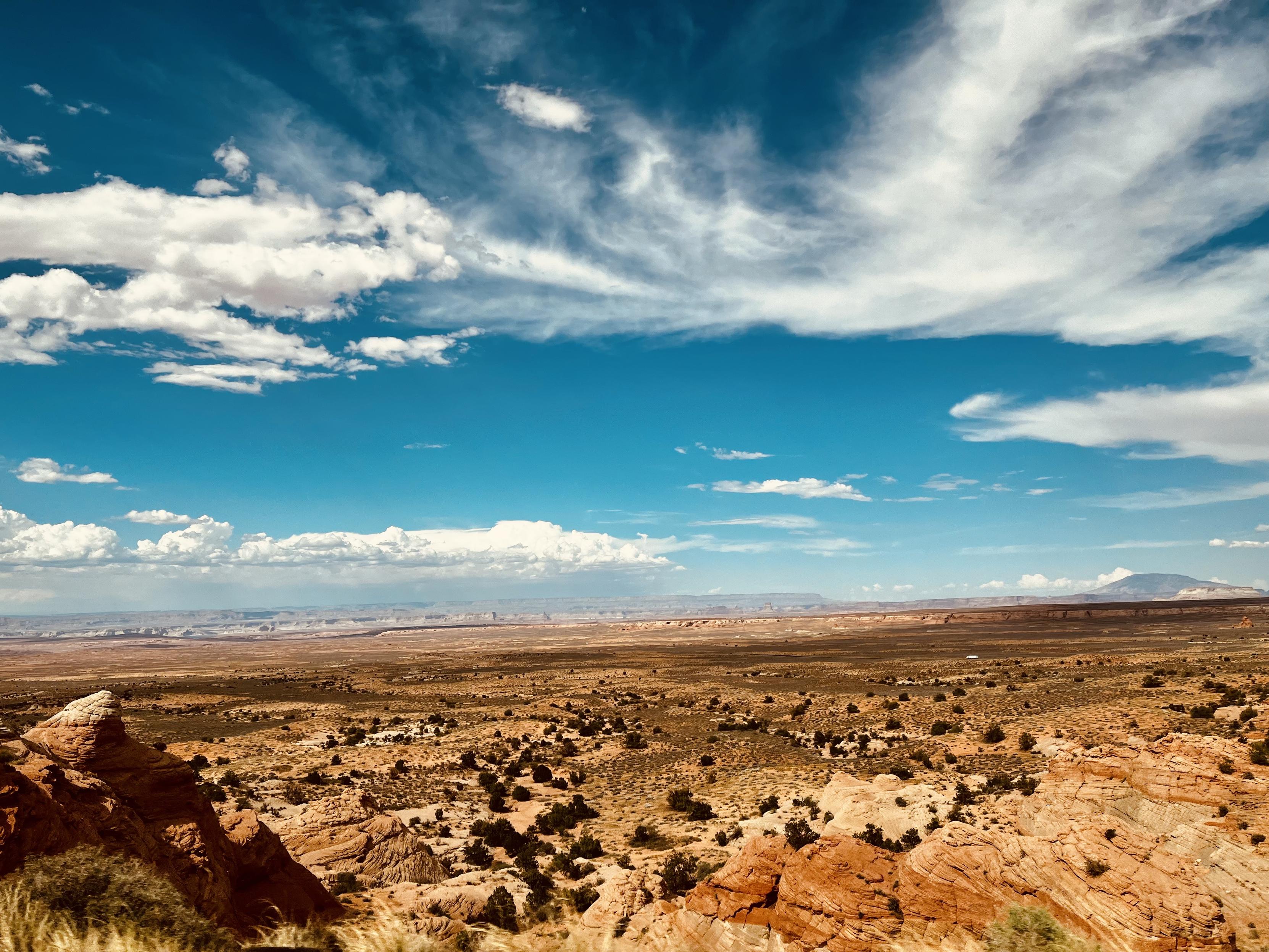 A color, wide-angle, landscape photograph capturing the vast, arid terrain of Coconino County, Arizona, under a brilliant blue afternoon sky. The foreground is dominated by rugged, reddish-brown rock formations and dry scrub brush. The closest rocks, particularly on the lower left, show sharp, angular edges and deep shadows that emphasize their texture. Scattered across the mid-ground, the sparse vegetation appears dark against the light-brown, sandy soil, creating a speckled pattern that stretches toward the horizon. The middle and background transition into an expansive, flat desert valley that extends towards distant mesas and plateaus. They appear hazy and layered in shades of blue and purple from the distance. The sky occupies roughly the top half of the frame, and scattered, wispy cirrus and cumulus clouds that form dynamic horizontal bands. A faint, low butte is visible on the far right horizon.