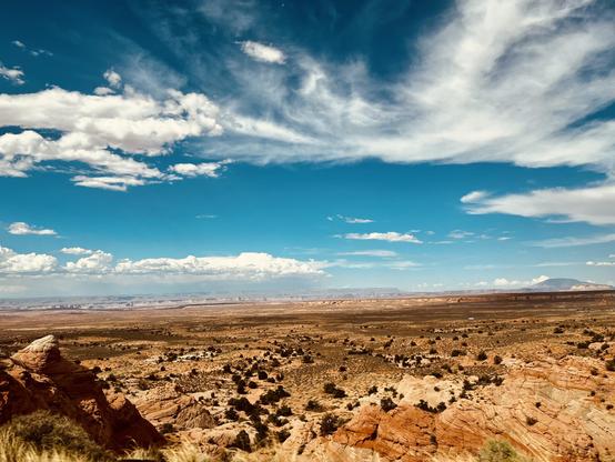 A color, wide-angle, landscape photograph capturing the vast, arid terrain of Coconino County, Arizona, under a brilliant blue afternoon sky. The foreground is dominated by rugged, reddish-brown rock formations and dry scrub brush. The closest rocks, particularly on the lower left, show sharp, angular edges and deep shadows that emphasize their texture. Scattered across the mid-ground, the sparse vegetation appears dark against the light-brown, sandy soil, creating a speckled pattern that stretches toward the horizon. The middle and background transition into an expansive, flat desert valley that extends towards distant mesas and plateaus. They appear hazy and layered in shades of blue and purple from the distance. The sky occupies roughly the top half of the frame, and scattered, wispy cirrus and cumulus clouds that form dynamic horizontal bands. A faint, low butte is visible on the far right horizon.