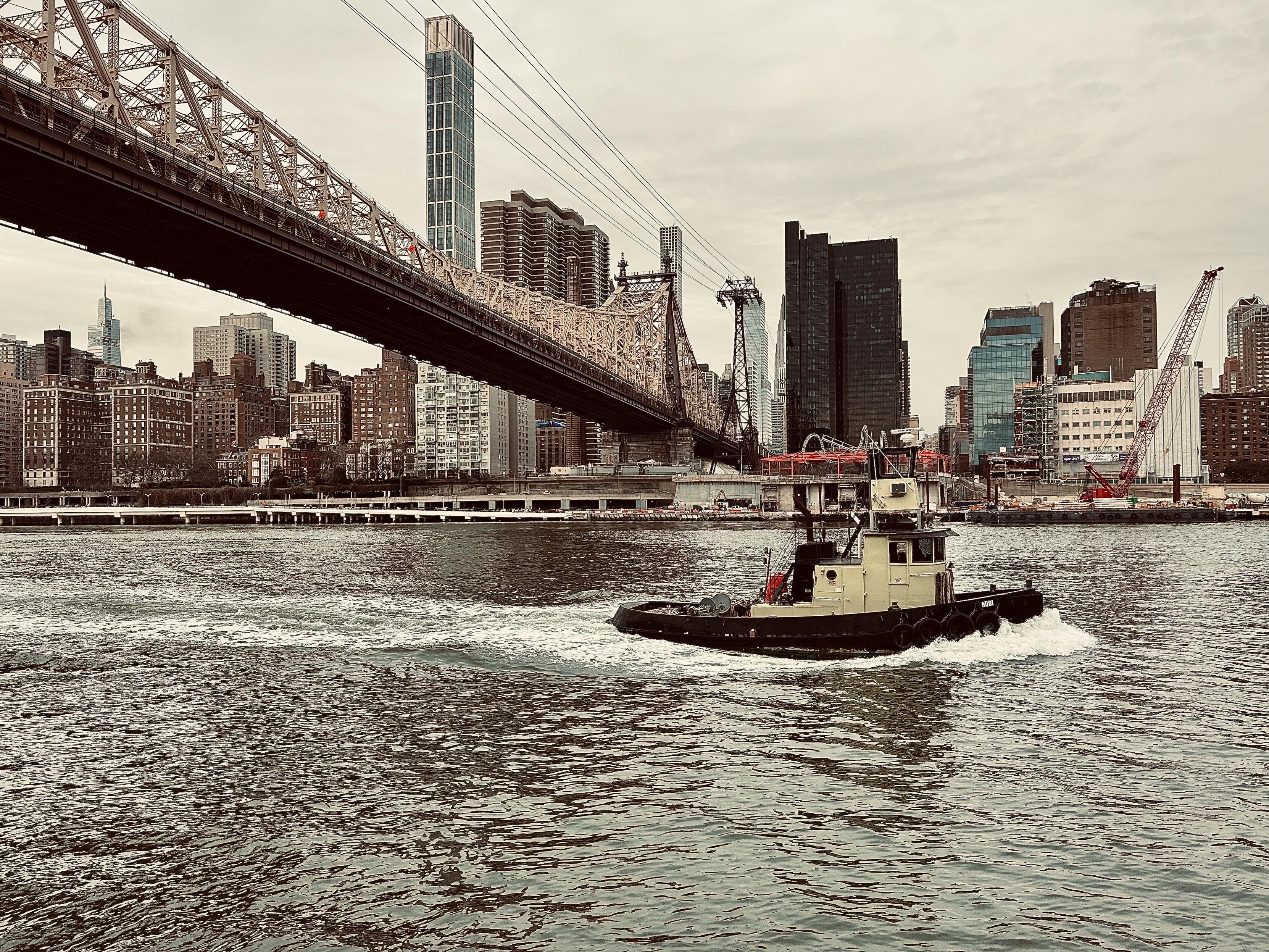 A color, wide-angle photograph that captures a scene on the East River in New York City. The image is desaturated, taken on a cloudy winter afternoon. The primary subject is a small, yellow and black color tugboat moving quickly across the foreground, creating a distinct white wake behind it on the choppy dark water. The tugboat is centered slightly to the right of the frame. Dominating the upper-left and center of the image is the massive, truss-supported Queensboro Bridge, which spans the river diagonally from the upper-left corner toward the center-right background. The background is a dense skyline of Midtown Manhattan, viewed from the perspective of Roosevelt Island. The buildings vary in height and style, featuring older brick apartment complexes on the far left, transitioning to modern glass and concrete skyscrapers in the center and right. There is active construction visible on the right, indicated by a large red and white construction crane.