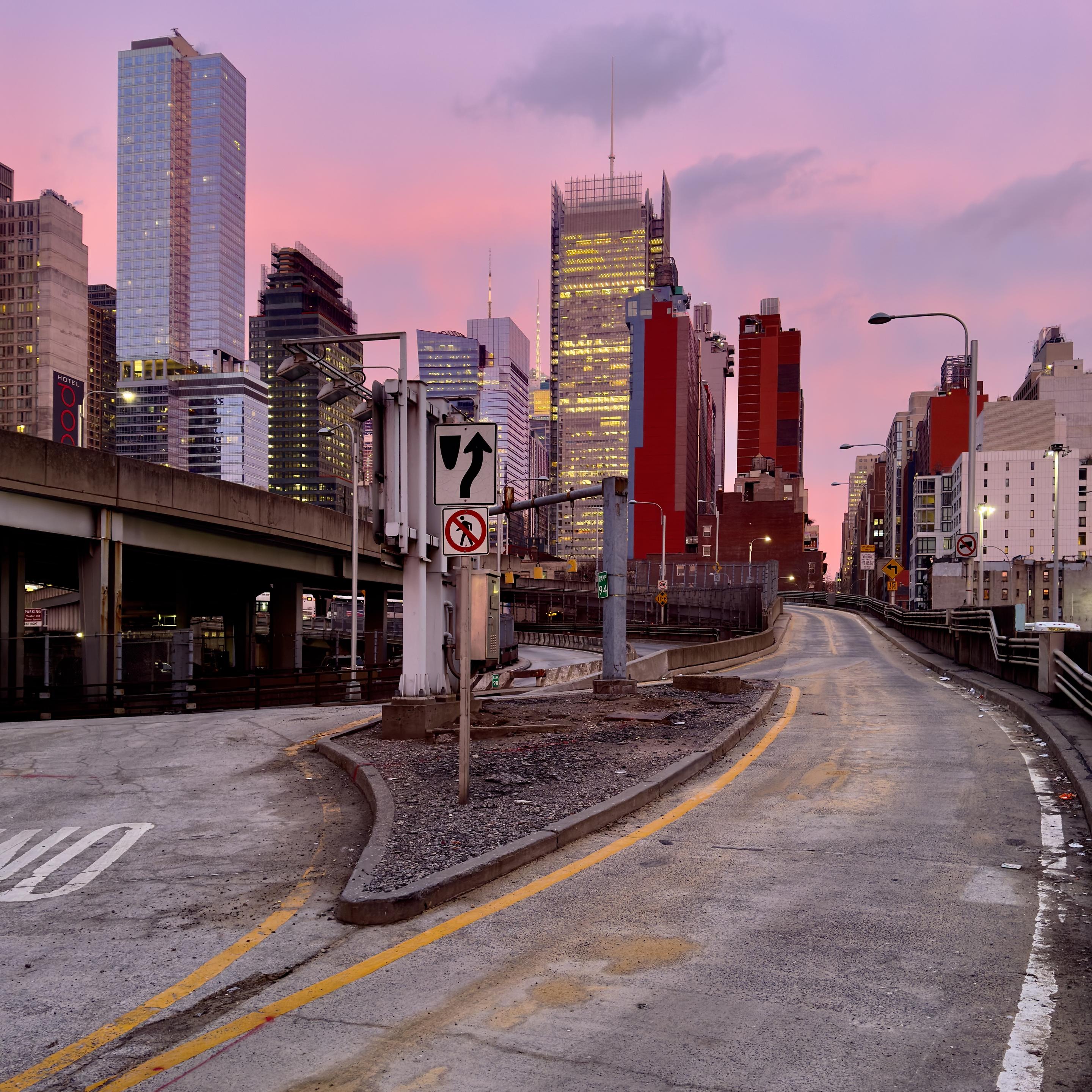A color photograph taken along 10th Avenue facing north from the Lincoln Tunnel Expressway at sunset. The central focus is a bifurcating, elevated asphalt roadway. The foreground features a triangular median curb covered in gravel and dirt, dividing the road. Attached to a utility pole on this median is a traffic sign. The roadway itself is worn, with yellow dashed and solid lines marking the lanes. To the left, a concrete elevated highway ramp, supported by columns, runs parallel to the main road. The background is dominated by the city skyline, vibrant pink and magenta hues of the sunset sky, which casts a soft, warm light over the scene. The New York Times Tower is in the center of the frame. Other skyscrapers of varying heights and architectural styles fill the frame, many with illuminated windows.