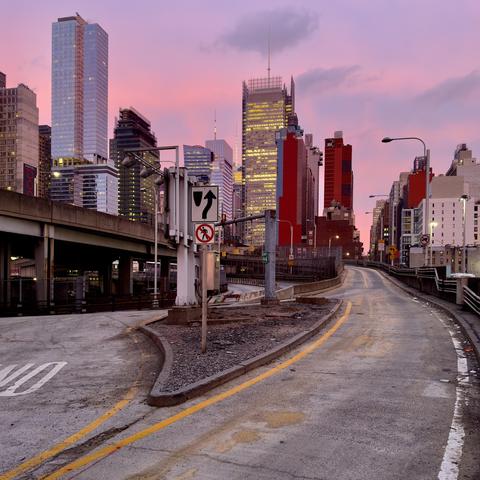 A color photograph taken along 10th Avenue facing north from the Lincoln Tunnel Expressway at sunset. The central focus is a bifurcating, elevated asphalt roadway. The foreground features a triangular median curb covered in gravel and dirt, dividing the road. Attached to a utility pole on this median is a traffic sign. The roadway itself is worn, with yellow dashed and solid lines marking the lanes. To the left, a concrete elevated highway ramp, supported by columns, runs parallel to the main road. The background is dominated by the city skyline, vibrant pink and magenta hues of the sunset sky, which casts a soft, warm light over the scene. The New York Times Tower is in the center of the frame. Other skyscrapers of varying heights and architectural styles fill the frame, many with illuminated windows.
