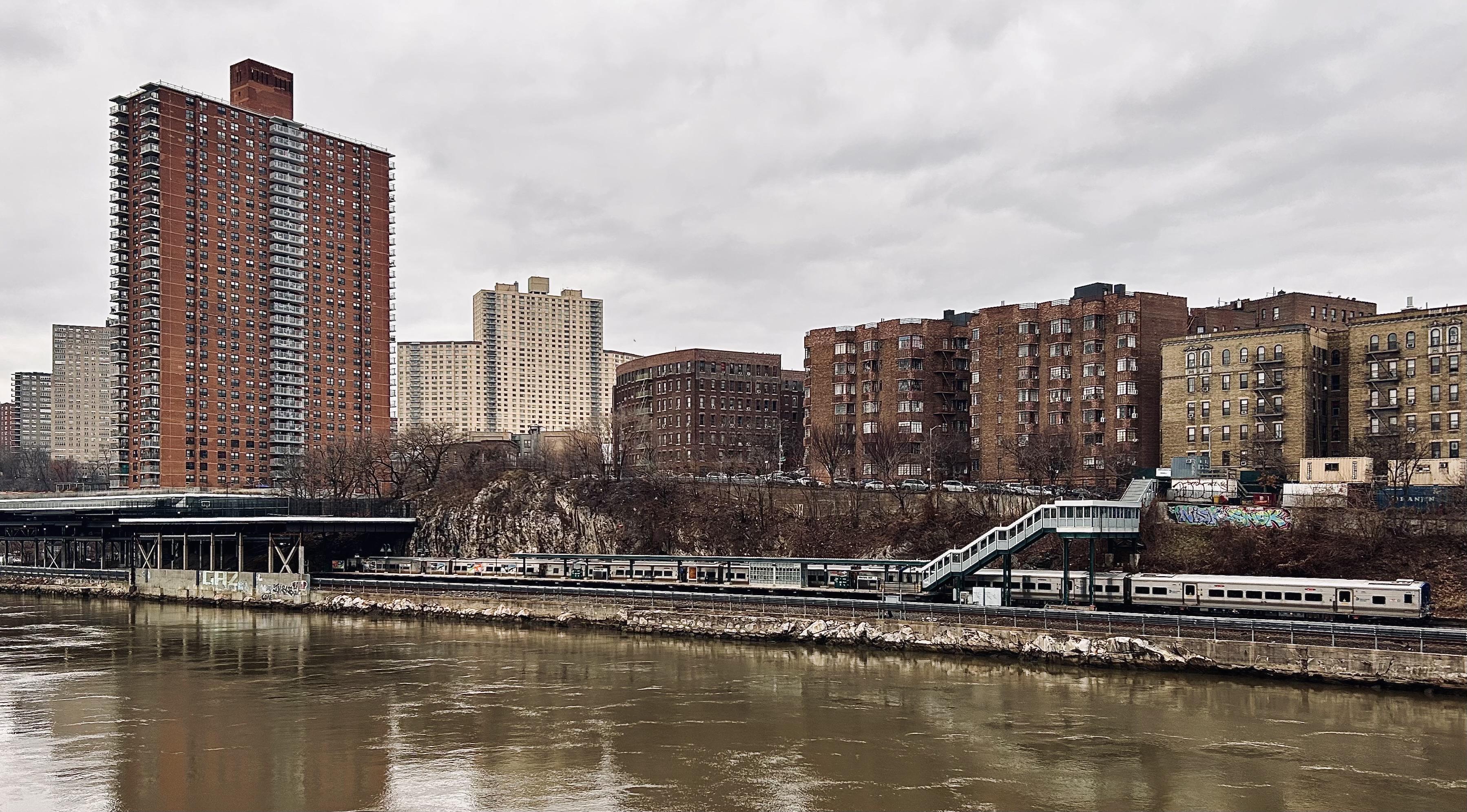A color, wide-angle photograph in landscape orientation that captures a gloomy, overcast day along the Harlem River, looking toward the Marble Hill neighborhood of Manhattan. In the foreground, the murky, brownish-green water of the river ripples slightly, reflecting the muted light of the gray sky. On the opposite bank, a Metro-North train station sits at the base of a rocky, dirt-covered hill. A long, silver commuter train is stopped at the platform, with a white pedestrian overpass bridge featuring green accents and glass windows arching over the tracks. Sparse, leafless brown trees and patches of dry brush line the hillside behind the station, where a few hints of colorful graffiti are visible on concrete retaining walls. The background is dominated by a dense urban skyline of varying architectural styles. To the left, a prominent, tall rectangular apartment building made of reddish-brown brick stands out against the sky, featuring rows of small white balconies. To its right, several other large, sandy-colored high-rise complexes rise further back. On the right side of the frame, a row of medium-rise, brick apartment buildings sits atop the ridge, characterized by their uniform fire escapes and flat roofs.
