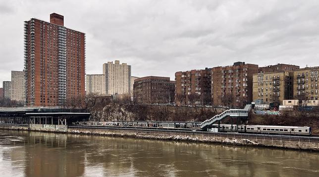 A color, wide-angle photograph in landscape orientation that captures a gloomy, overcast day along the Harlem River, looking toward the Marble Hill neighborhood of Manhattan. In the foreground, the murky, brownish-green water of the river ripples slightly, reflecting the muted light of the gray sky. On the opposite bank, a Metro-North train station sits at the base of a rocky, dirt-covered hill. A long, silver commuter train is stopped at the platform, with a white pedestrian overpass bridge featuring green accents and glass windows arching over the tracks. Sparse, leafless brown trees and patches of dry brush line the hillside behind the station, where a few hints of colorful graffiti are visible on concrete retaining walls. The background is dominated by a dense urban skyline of varying architectural styles. To the left, a prominent, tall rectangular apartment building made of reddish-brown brick stands out against the sky, featuring rows of small white balconies. To its right, several other large, sandy-colored high-rise complexes rise further back. On the right side of the frame, a row of medium-rise, brick apartment buildings sits atop the ridge, characterized by their uniform fire escapes and flat roofs.