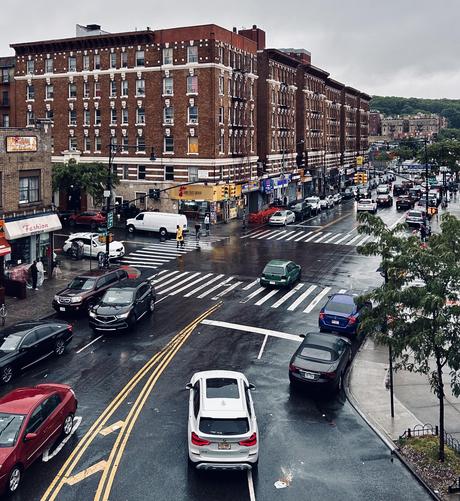 A color photograph from an elevated view captures a busy, rain-slicked street in the Inwood neighborhood of Manhattan on an overcast day morning. Large, multi-story pre-war apartment buildings constructed of red brick with ornate white window trim line the wide city streets. The asphalt is dark and reflective from the rain, showing the blurred reflections of traffic lights and vehicle headlights. A mix of colorful cars, including a prominent white SUV in the foreground and a green hatchback mid-intersection, navigate the wet roads, while white painted crosswalks and double yellow lines create a geometric pattern across the pavement. On the street level, various local storefronts are visible, including one with a bright yellow sign for a fried chicken restaurant and another called "Lara Fashion" with a white awning. A few pedestrians, one wearing a bright yellow raincoat, are seen crossing the street or standing on the sidewalks. Sparse green trees with thin branches provide a touch of nature against the dense urban landscape. In the far distance, the gray sky meets the hazy, green foliage of a nearby hilly city park.