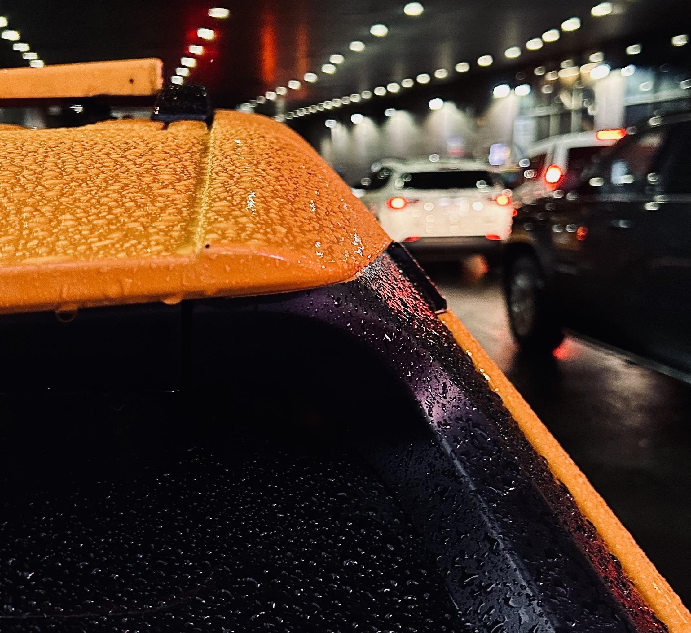 A color photograph in square aspect ratio. A close-up shot that focuses on the rear roofline of a vibrant yellow taxi, covered in thousands of tiny, glistening raindrops. The yellow surface is textured with beads of water that catch the overhead lights, while a black trim piece below it creates a sharp contrast, also saturated with fine mist. To the left, the edge of the taxi roof sign is visible, partially obscured by the angle. The perspective is low and tight, emphasizing the physical texture of the wet car surface against the dark, moody atmosphere of the transit hub. In the blurred background, the street becomes a tunnel beneath the Port Authority terminal. A series of glowing circular lights line the ceiling and recede into the distance. The wet pavement reflects the red and white glow of brake lights and headlights from a line of idling cars. A white SUV and a dark van are visible in soft focus.