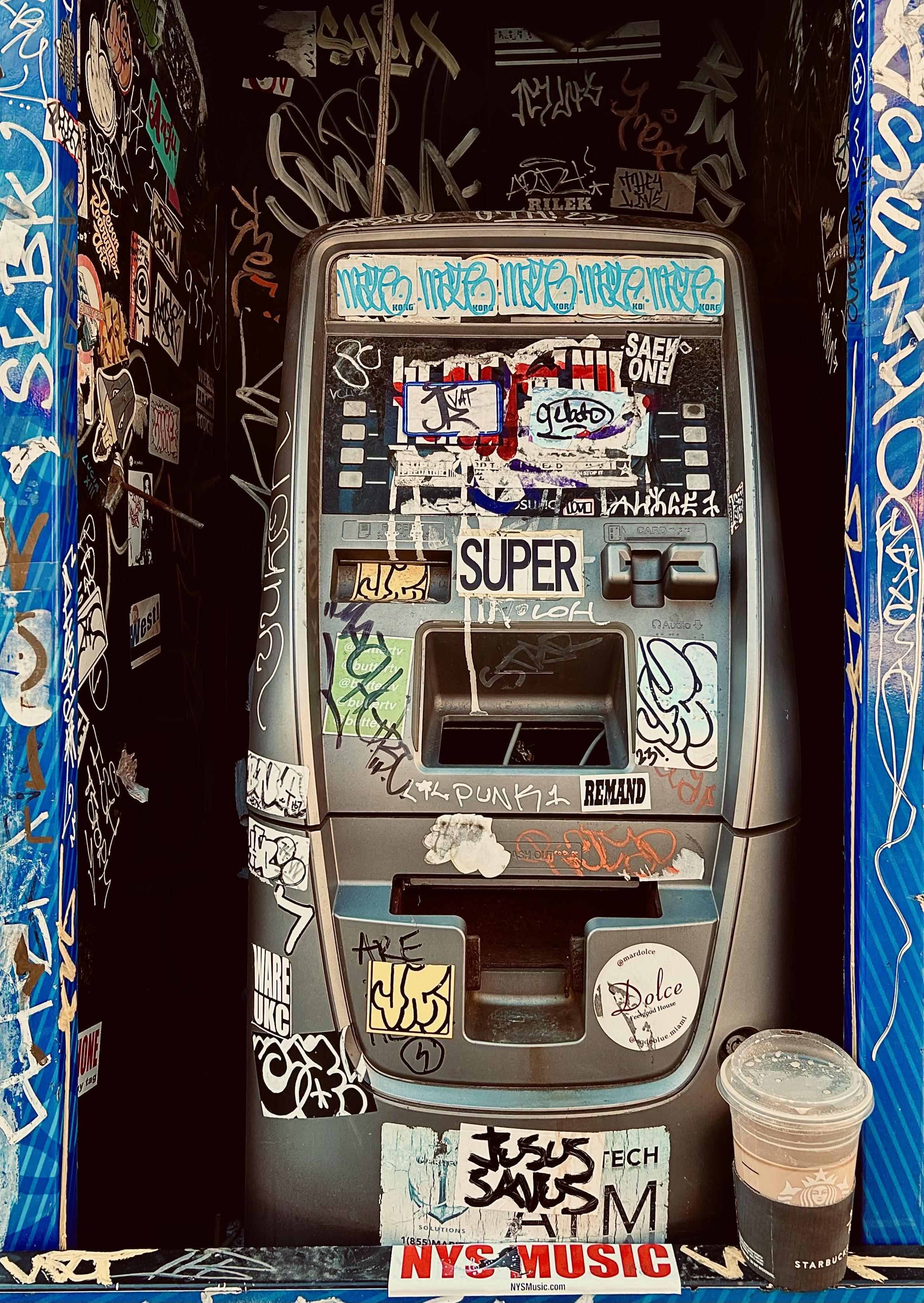 A color photograph in portrait orientation captures a decommissioned silver ATM housed within a narrow, dark alcove along Eighth Avenue in the Chelsea neighborhood of Manhattam on a partly cloudy afternoon. The machine and its surrounding walls are almost entirely covered in a dense layer of street art, including colorful vinyl stickers, hand-drawn art, and scrawled graffiti tags in white, black, and blue ink. A prominent "SUPER" sticker is centered above the keypad area, while a "JESUS SAVES" tag is visible near the bottom. The ATM itself is weathered, with its screen obscured by stickers and its card slot and cash dispenser are empty and hollow, giving the machine a skeletal appearance. The interior walls of the booth are black, serving as a dark canvas for numerous white paint-pen signatures and overlapping stickers. To the bottom, left, and right a blue-patterned frame borders the booth, and a half-full Starbucks iced coffee cup with a brown sleeve sits on the ledge in front of the machine. At the very bottom edge of the frame, a red and white sticker reads "NYS MUSIC." The lighting is somewhat dim, warm, and moody.