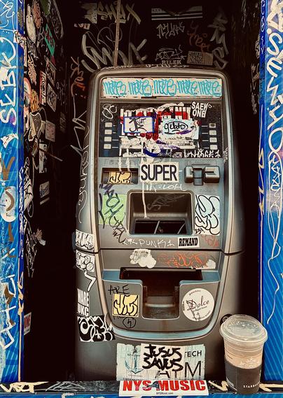 A color photograph in portrait orientation captures a decommissioned silver ATM housed within a narrow, dark alcove along Eighth Avenue in the Chelsea neighborhood of Manhattam on a partly cloudy afternoon. The machine and its surrounding walls are almost entirely covered in a dense layer of street art, including colorful vinyl stickers, hand-drawn art, and scrawled graffiti tags in white, black, and blue ink. A prominent "SUPER" sticker is centered above the keypad area, while a "JESUS SAVES" tag is visible near the bottom. The ATM itself is weathered, with its screen obscured by stickers and its card slot and cash dispenser are empty and hollow, giving the machine a skeletal appearance. The interior walls of the booth are black, serving as a dark canvas for numerous white paint-pen signatures and overlapping stickers. To the bottom, left, and right a blue-patterned frame borders the booth, and a half-full Starbucks iced coffee cup with a brown sleeve sits on the ledge in front of the machine. At the very bottom edge of the frame, a red and white sticker reads "NYS MUSIC." The lighting is somewhat dim, warm, and moody.