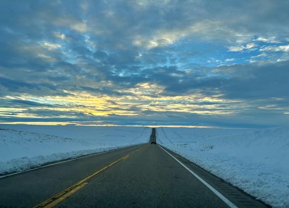A color photograph taken along interstate 70 somewhere west of Quinter, Kansas. It’s late afternoon on a winter day and the sun is setting over a vast flat landscape covered in snow. Many clouds fill the light blue sky. The sun is shining through spots to vast yellow and darker blue color across the sky. The highway is two lanes and in the distance a semi truck is seen driving away from the camera. The overall mood of the scene is cool and calm.