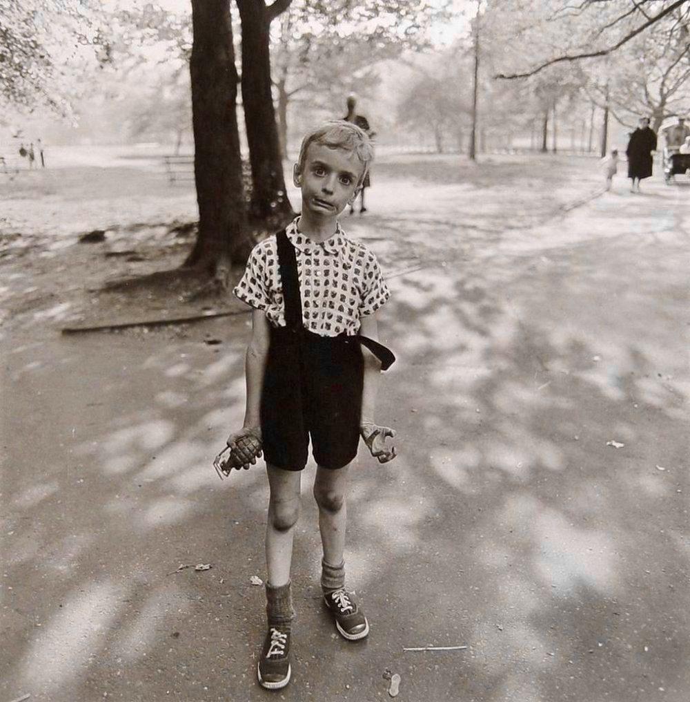 A black-and-white full-length portrait captures a young, slender boy standing on a paved path in Central Park, New York City, in the summer of 1962. He wears a patterned short-sleeved button-down shirt under dark overalls, with one strap hanging loosely off his shoulder. His expression is highly animated and tense; his eyes are wide and bulging, his brow is furrowed, and his lips are pulled into a stiff, manic grimace. His thin arms hang rigidly at his sides, with his right hand tightly clutching a realistic toy hand grenade and his left hand curled into a stiff, claw-like gesture. The background is softly blurred, showing the sunlight and shadows of large trees. In the distance, a few indistinct figures of people walking and a baby carriage. An out of focus person is standing a short distance behind the boy next to a tree. The lighting is bright but filtered through the canopy, casting intricate leaf shadows across the ground and the boy’s clothing. Photograph taken by Diane Arbus.