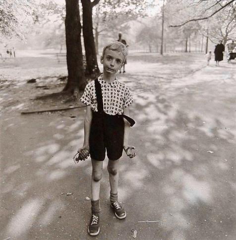 A black-and-white full-length portrait captures a young, slender boy standing on a paved path in Central Park, New York City, in the summer of 1962. He wears a patterned short-sleeved button-down shirt under dark overalls, with one strap hanging loosely off his shoulder. His expression is highly animated and tense; his eyes are wide and bulging, his brow is furrowed, and his lips are pulled into a stiff, manic grimace. His thin arms hang rigidly at his sides, with his right hand tightly clutching a realistic toy hand grenade and his left hand curled into a stiff, claw-like gesture. The background is softly blurred, showing the sunlight and shadows of large trees. In the distance, a few indistinct figures of people walking and a baby carriage. An out of focus person is standing a short distance behind the boy next to a tree. The lighting is bright but filtered through the canopy, casting intricate leaf shadows across the ground and the boy’s clothing. Photograph taken by Diane Arbus.