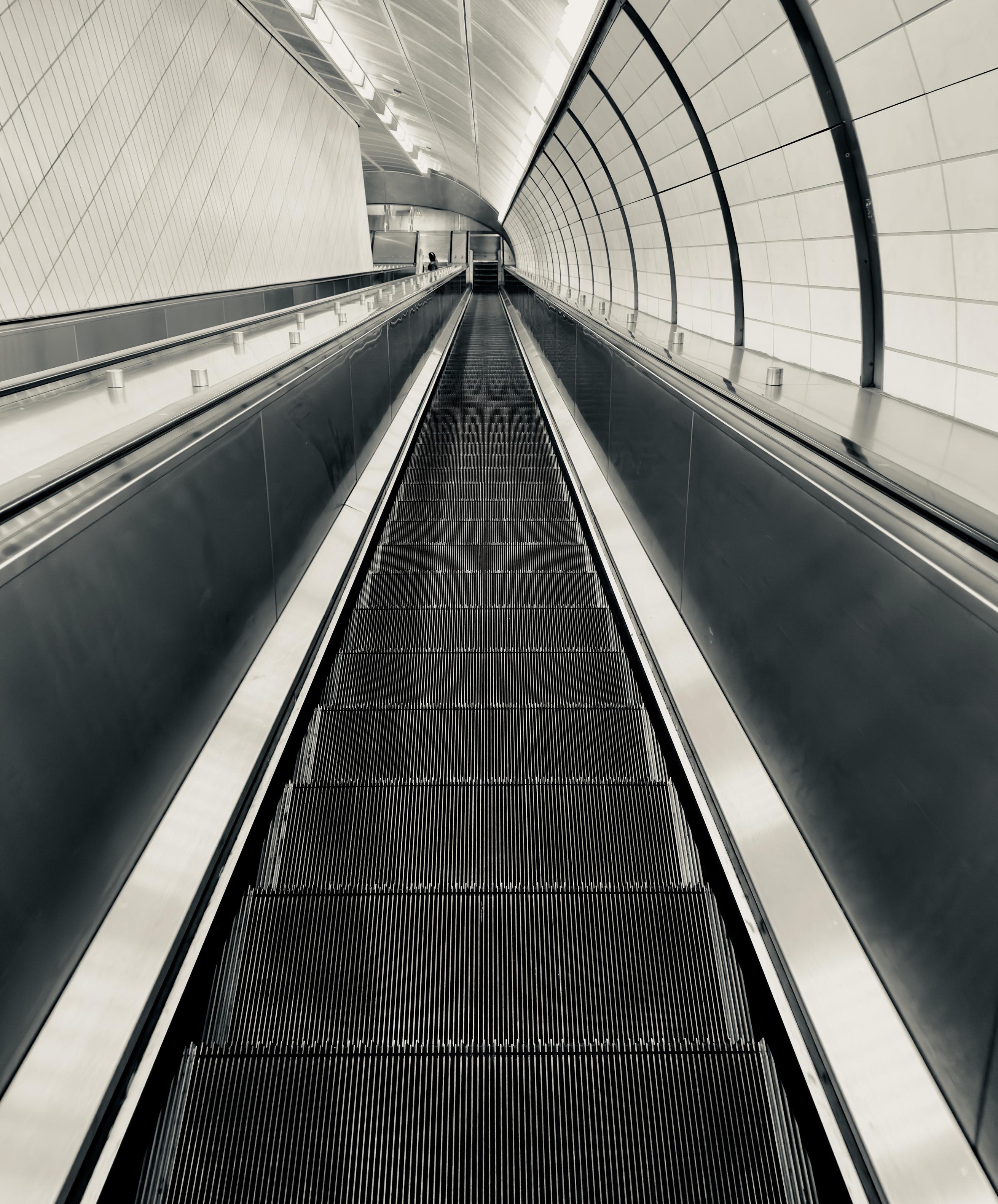 A silvertone, high-angle photograph of a steep, seemingly endless descent down a modern escalator at the Hudson Yards subway station in Manhattan. The composition is symmetrical, with the vertical lines of the escalator steps drawing the eye toward a distant, vanishing point at the bottom of the incline. To the right, a curved wall is lined with large, rectangular white tiles, while the left side features a sleek, metallic partition and a textured, diagonal-patterned wall. The monochromatic palette emphasizes the interplay of light and shadow, highlighting the polished surfaces of the handrails and the repetitive, industrial texture of the escalator treads. The perspective creates a sense of immense depth and scale. Recessed lighting along the vaulted ceiling casts a soft, rhythmic glow that follows the curvature of the tunnel, reflecting off the dark, metallic side panels of the escalator. In the far distance, a lone person can be seen near the bottom, providing a sense of human scale against the vast, cavernous architecture. The overall atmosphere is one of quiet, futuristic minimalism, focusing on the clean geometric lines.