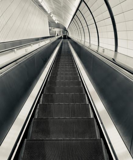 A silvertone, high-angle photograph of a steep, seemingly endless descent down a modern escalator at the Hudson Yards subway station in Manhattan. The composition is symmetrical, with the vertical lines of the escalator steps drawing the eye toward a distant, vanishing point at the bottom of the incline. To the right, a curved wall is lined with large, rectangular white tiles, while the left side features a sleek, metallic partition and a textured, diagonal-patterned wall. The monochromatic palette emphasizes the interplay of light and shadow, highlighting the polished surfaces of the handrails and the repetitive, industrial texture of the escalator treads. The perspective creates a sense of immense depth and scale. Recessed lighting along the vaulted ceiling casts a soft, rhythmic glow that follows the curvature of the tunnel, reflecting off the dark, metallic side panels of the escalator. In the far distance, a lone person can be seen near the bottom, providing a sense of human scale against the vast, cavernous architecture. The overall atmosphere is one of quiet, futuristic minimalism, focusing on the clean geometric lines.