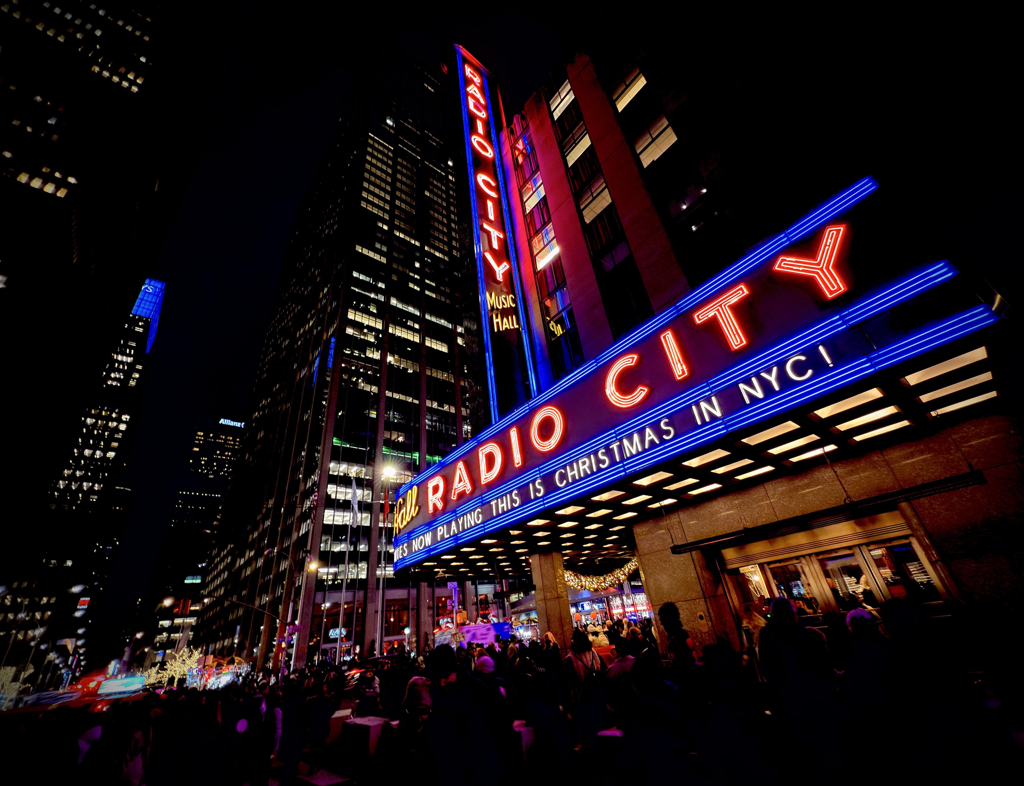 A color, low-angle photograph in landscape orientation taken at night of the iconic facade of Radio City Music Hall in New York City, illuminated with vibrant neon lights. The Art Deco building's marquee stretches across the frame, glowing with bright red letters outlined in blue neon that spell out "RADIO CITY." Below the main name, a smaller electronic ticker displays the message, "NOW PLAYING THIS IS CHRISTMAS IN NYC!" A towering vertical sign climbs the side of the dark skyscraper, repeating the venue's name in stacked, glowing red letters. The perspective is tilted slightly, creating a dynamic sense of height and energy against the deep black night sky. The street level is filled with a dense, silhouetted crowd of many people gathered under the warm, golden glow of the marquee’s underside. In the background, the city's urban landscape is visible through the cool blue lights of neighboring skyscrapers and distant holiday decorations, including a glimpse of a lit Christmas tree.