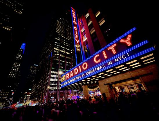 A color, low-angle photograph in landscape orientation taken at night of the iconic facade of Radio City Music Hall in New York City, illuminated with vibrant neon lights. The Art Deco building's marquee stretches across the frame, glowing with bright red letters outlined in blue neon that spell out "RADIO CITY." Below the main name, a smaller electronic ticker displays the message, "NOW PLAYING THIS IS CHRISTMAS IN NYC!" A towering vertical sign climbs the side of the dark skyscraper, repeating the venue's name in stacked, glowing red letters. The perspective is tilted slightly, creating a dynamic sense of height and energy against the deep black night sky. The street level is filled with a dense, silhouetted crowd of many people gathered under the warm, golden glow of the marquee’s underside. In the background, the city's urban landscape is visible through the cool blue lights of neighboring skyscrapers and distant holiday decorations, including a glimpse of a lit Christmas tree.