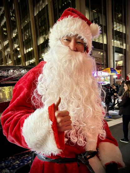 A color, medium-angle portrait photograph that captures a person dressed as Santa Claus standing outdoors at night in the Theater District neighborhood of Manhattan at night. The individual wears a vibrant red Santa suit with plush white faux-fur trim along the hood and sleeves. A long, curly white synthetic beard and mustache cover most of their face, though their smiling eyes and the bridge of their nose remain visible. They are giving a cheerful thumbs-up toward the camera with their right hand, which is framed by the red and white sleeve of the costume. The background features towering, illuminated office buildings with rows of rectangular windows glowing against the dark sky. To the right, the blurred activity of a city street is visible, including a food vendor’s cart with a colorful yellow and blue umbrella and several pedestrians in winter attire walking by.