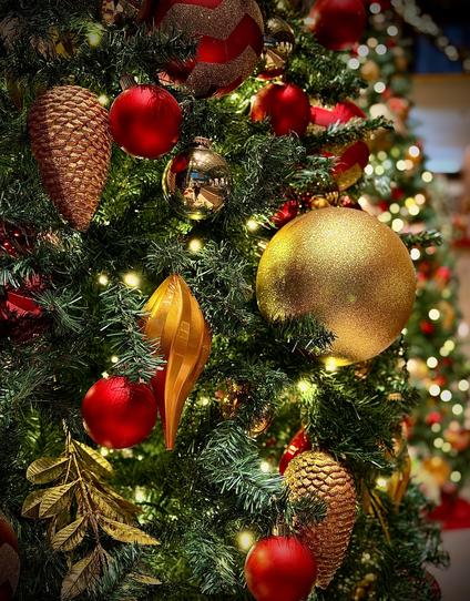 A color, close-up photograph in portrait orientation that captures an artificial tree that has been decorated for Christmas. The dense, uniform green needles are adorned with a variety of festive lights and ornaments. The primary color palette consists of shimmering golds and rich deep reds. Key decorations include matte and metallic red spherical baubles, a large textured gold glitter ball, and two gold and copper colored pinecone ornaments with intricate scale patterns. A prominent, elongated gold teardrop ornament with vertical ridges hangs near the center, while several golden artificial leaves are tucked into the branches. Small, warm white fairy lights are scattered throughout, casting a soft glow on the ornaments. The composition uses a shallow depth of field, keeping the ornaments in the immediate foreground in sharp focus while the background softly blurs into a bokeh of golden lights and additional tree branches. One polished gold ornament in the upper center acts as a mirror, reflecting a distorted view of the brightly lit hotel lobby and I am partially visible taking the photo wearing blue jeans, black dress shoes, a black hoodie, and winter cap. Both of my hands are holding onto a silver iPhone 17 Pro Max. The lighting is warm and directional, creating highlights on the smooth surfaces of the red baubles and a sparkling texture on the glittered elements.