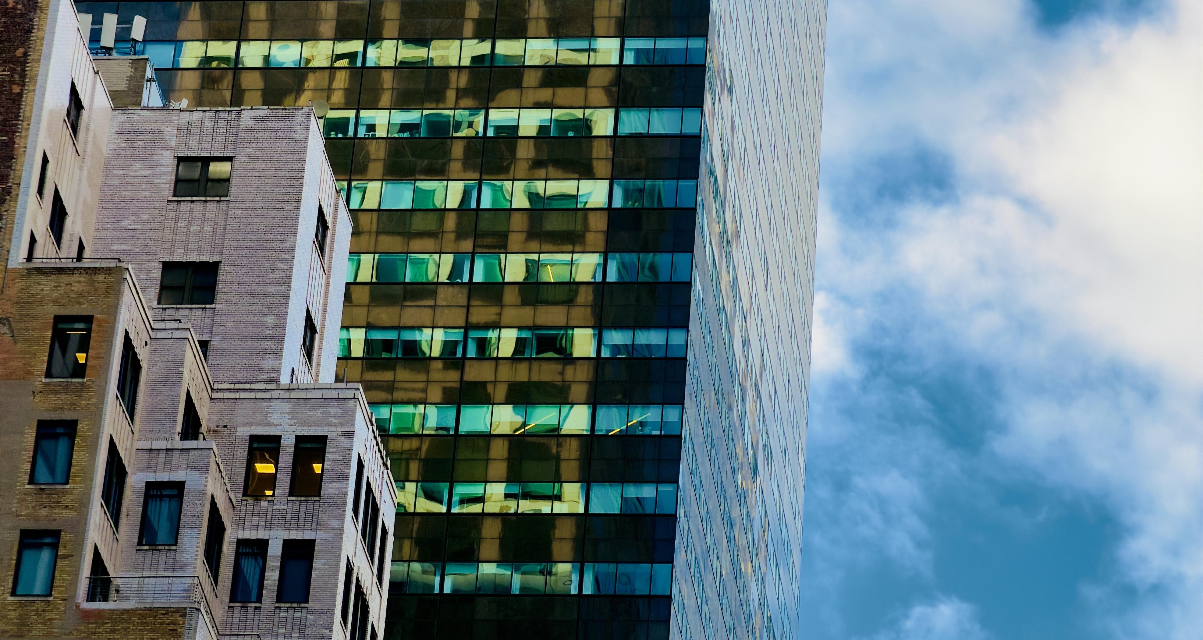 A color photograph in landscape orientation that captures an architectural close-up of a vertical slice of Manhattan’s skyline against a medium blue sky filled with scattered white clouds. In the foreground on the left, an older masonry building features a stepped, wedding-cake style facade with light-colored brick and a series of dark-framed windows, some of which are illuminated from within by a warm golden glow. This traditional structure contrasts sharply with the massive glass skyscraper behind it, which dominates the center and right of the frame. The modern tower’s reflective surface is divided into a grid of windows that catch the intense light of the golden hour, creating a rhythmic pattern of amber, turquoise, and deep shadow. The composition emphasizes the density and verticality of the city through a tight crop that eliminates the street level, focusing instead on the intersection of different architectural eras. The golden hour light casts a brilliant sheen on the glass panels, mirroring the sky and nearby buildings in distorted, shimmering patches of color. Towards the far right, the northern edge of the building is visible, its surface reflecting the cool tones of the sky.
