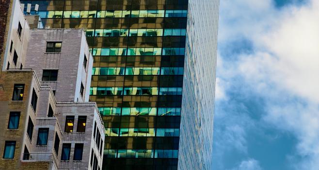 A color photograph in landscape orientation that captures an architectural close-up of a vertical slice of Manhattan’s skyline against a medium blue sky filled with scattered white clouds. In the foreground on the left, an older masonry building features a stepped, wedding-cake style facade with light-colored brick and a series of dark-framed windows, some of which are illuminated from within by a warm golden glow. This traditional structure contrasts sharply with the massive glass skyscraper behind it, which dominates the center and right of the frame. The modern tower’s reflective surface is divided into a grid of windows that catch the intense light of the golden hour, creating a rhythmic pattern of amber, turquoise, and deep shadow. The composition emphasizes the density and verticality of the city through a tight crop that eliminates the street level, focusing instead on the intersection of different architectural eras. The golden hour light casts a brilliant sheen on the glass panels, mirroring the sky and nearby buildings in distorted, shimmering patches of color. Towards the far right, the northern edge of the building is visible, its surface reflecting the cool tones of the sky.