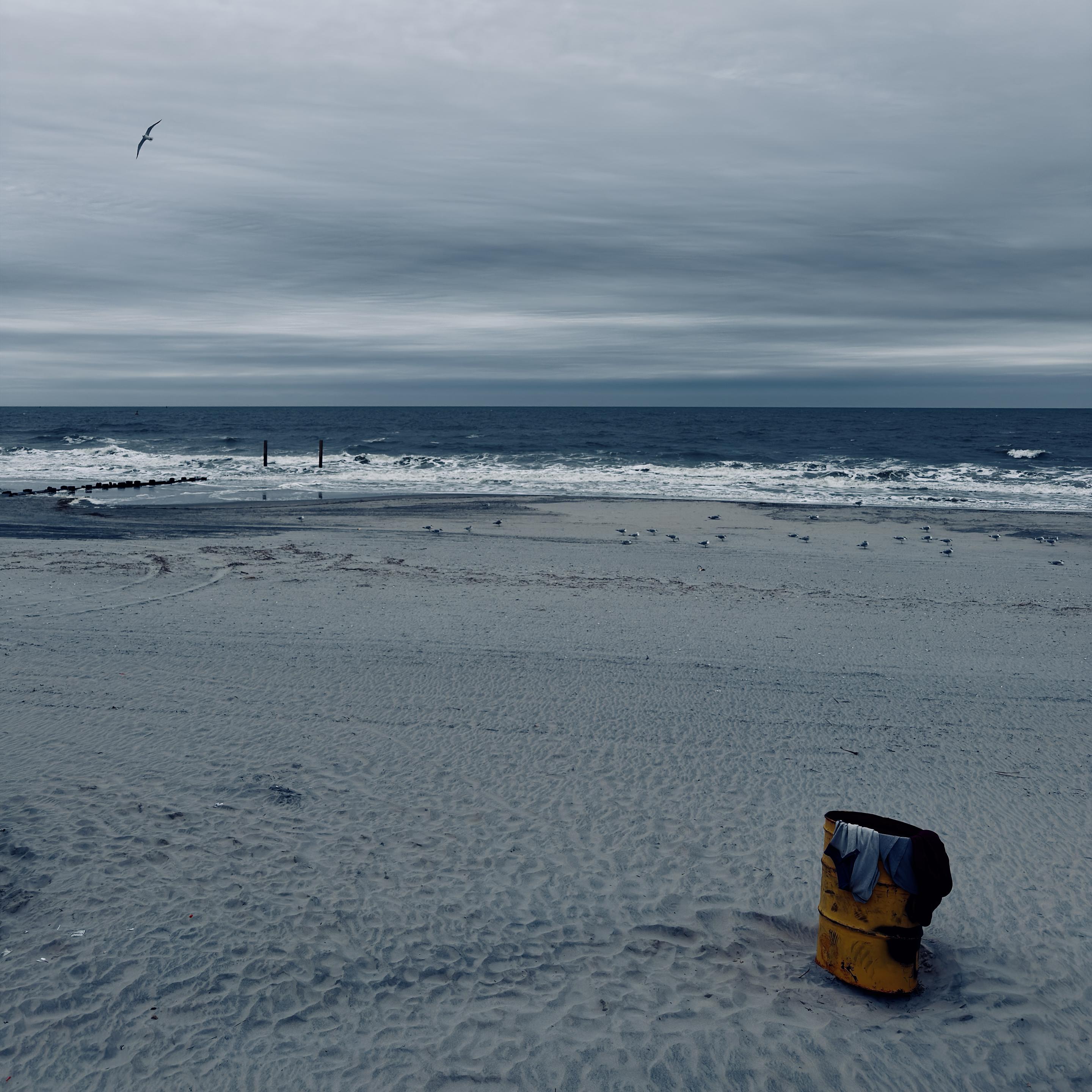 A color, medium-angle photograph in square aspect ratio. An eye-level shot captures a desaturated and overcast day at an Atlantic City beach. In the lower right foreground, a rusted, bright yellow metal trash barrel sits partially buried in the sand, with several dark and light-colored articles of clothing draped over its rim. The vast expanse of the beach is composed of pale, grey-toned sand textured with countless footprints and subtle ripples. In the mid-ground, a scattered flock of seagulls rests near the shoreline, while the dark blue ocean waves break into white foam against the coast. Two dark wooden pilings stand vertically in the shallow water, and a low rock jetty extends into the surf on the left. The upper half of the image is dominated by a heavy, moody sky filled with thick layers of horizontal grey clouds. A single seagull is captured in flight in the upper left, its wings spread against the overcast backdrop. The overall color palette is muted and cool, leaning into deep blues and slate greys, which makes the weathered yellow of the trash can stand out as the primary focal point. The horizon line is sharp and flat, separating the turbulent dark water from the expansive, gloomy sky, evoking a sense of quiet isolation on a cold coastal day.