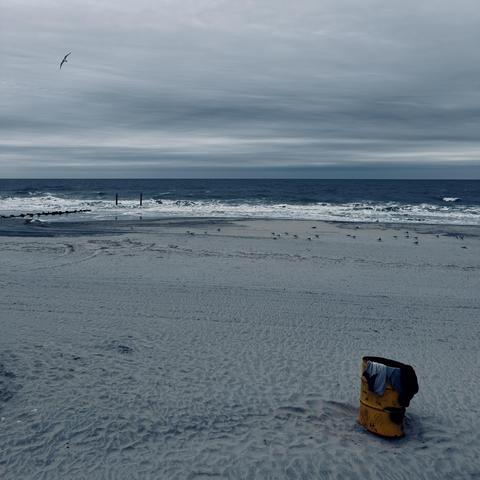 A color, medium-angle photograph in square aspect ratio. An eye-level shot captures a desaturated and overcast day at an Atlantic City beach. In the lower right foreground, a rusted, bright yellow metal trash barrel sits partially buried in the sand, with several dark and light-colored articles of clothing draped over its rim. The vast expanse of the beach is composed of pale, grey-toned sand textured with countless footprints and subtle ripples. In the mid-ground, a scattered flock of seagulls rests near the shoreline, while the dark blue ocean waves break into white foam against the coast. Two dark wooden pilings stand vertically in the shallow water, and a low rock jetty extends into the surf on the left. The upper half of the image is dominated by a heavy, moody sky filled with thick layers of horizontal grey clouds. A single seagull is captured in flight in the upper left, its wings spread against the overcast backdrop. The overall color palette is muted and cool, leaning into deep blues and slate greys, which makes the weathered yellow of the trash can stand out as the primary focal point. The horizon line is sharp and flat, separating the turbulent dark water from the expansive, gloomy sky, evoking a sense of quiet isolation on a cold coastal day.