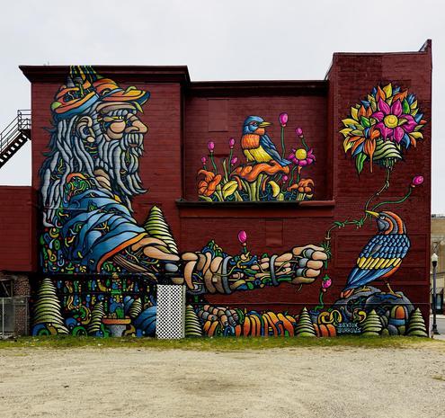 A color, medium-angle photograph in square aspect ratio that captures a vibrant, large-scale mural covering the multi-level dark red brick building. On the left, a profile of an elderly man with a long gray beard and a colorful, patterned hat gazes toward the right. His figure is composed of intricate, psychedelic swirls of blue, yellow, and green, resembling a mosaic. His arm extends across the center of the wall, where his hand is interacting with nature. At the bottom, a landscape features bright green conical trees and orange rounded shapes that mimic a forest floor. In the middle a small blue and orange bird sits perched. To the far right, a second bird with blue and yellow plumage stands on a rock, looking up at a giant, multi-colored flower. A small signature for "Denton Burrows" is visible near the bottom right corner, just above a patch of green grass and the gravel lot in the foreground. The overcast afternoon winter sky defines the horizon.