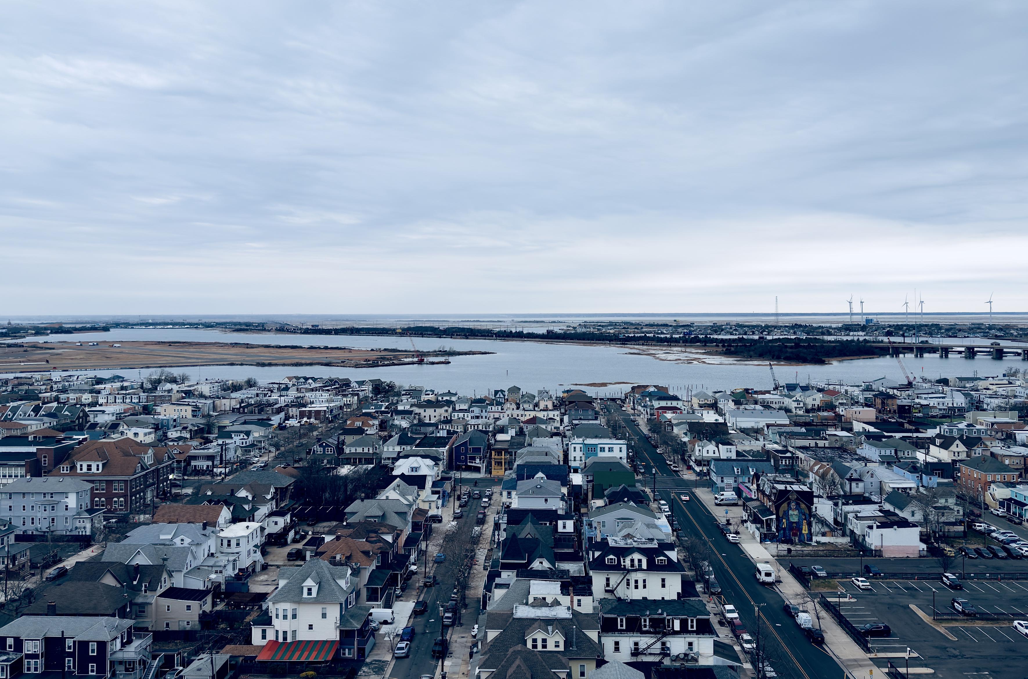 A color, wide-angle photograph in landscape orientation that captures an elevated, high-angle view of Atlantic City looking inland, away from the ocean, under a vast, overcast sky. The foreground and middle ground are densely packed with a mix of residential and small commercial buildings, featuring a variety of architectural styles including multi-story houses with gabled roofs, colorful siding, and brick exteriors. A straight, wide asphalt road cuts vertically through the right side of the frame, lined with parked cars and a few moving vehicles. To the far right, a large surface parking lot is visible, while a building with a vibrant, tall mural stands out among the more traditional structures. In the background, the urban landscape gives way to the waters of the interior thoroughfare and marshes. A long bridge spans across the water on the right side, leading toward the mainland. On the distant horizon a flat landscape is visible beneath the gray clouds, punctuated by a row of white wind turbines standing tall against the horizon line.