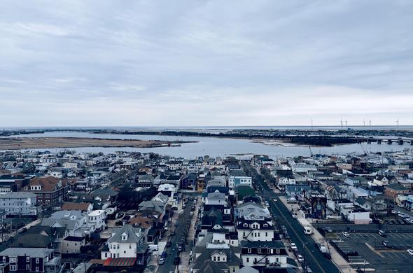 A color, wide-angle photograph in landscape orientation that captures an elevated, high-angle view of Atlantic City looking inland, away from the ocean, under a vast, overcast sky. The foreground and middle ground are densely packed with a mix of residential and small commercial buildings, featuring a variety of architectural styles including multi-story houses with gabled roofs, colorful siding, and brick exteriors. A straight, wide asphalt road cuts vertically through the right side of the frame, lined with parked cars and a few moving vehicles. To the far right, a large surface parking lot is visible, while a building with a vibrant, tall mural stands out among the more traditional structures. In the background, the urban landscape gives way to the waters of the interior thoroughfare and marshes. A long bridge spans across the water on the right side, leading toward the mainland. On the distant horizon a flat landscape is visible beneath the gray clouds, punctuated by a row of white wind turbines standing tall against the horizon line.